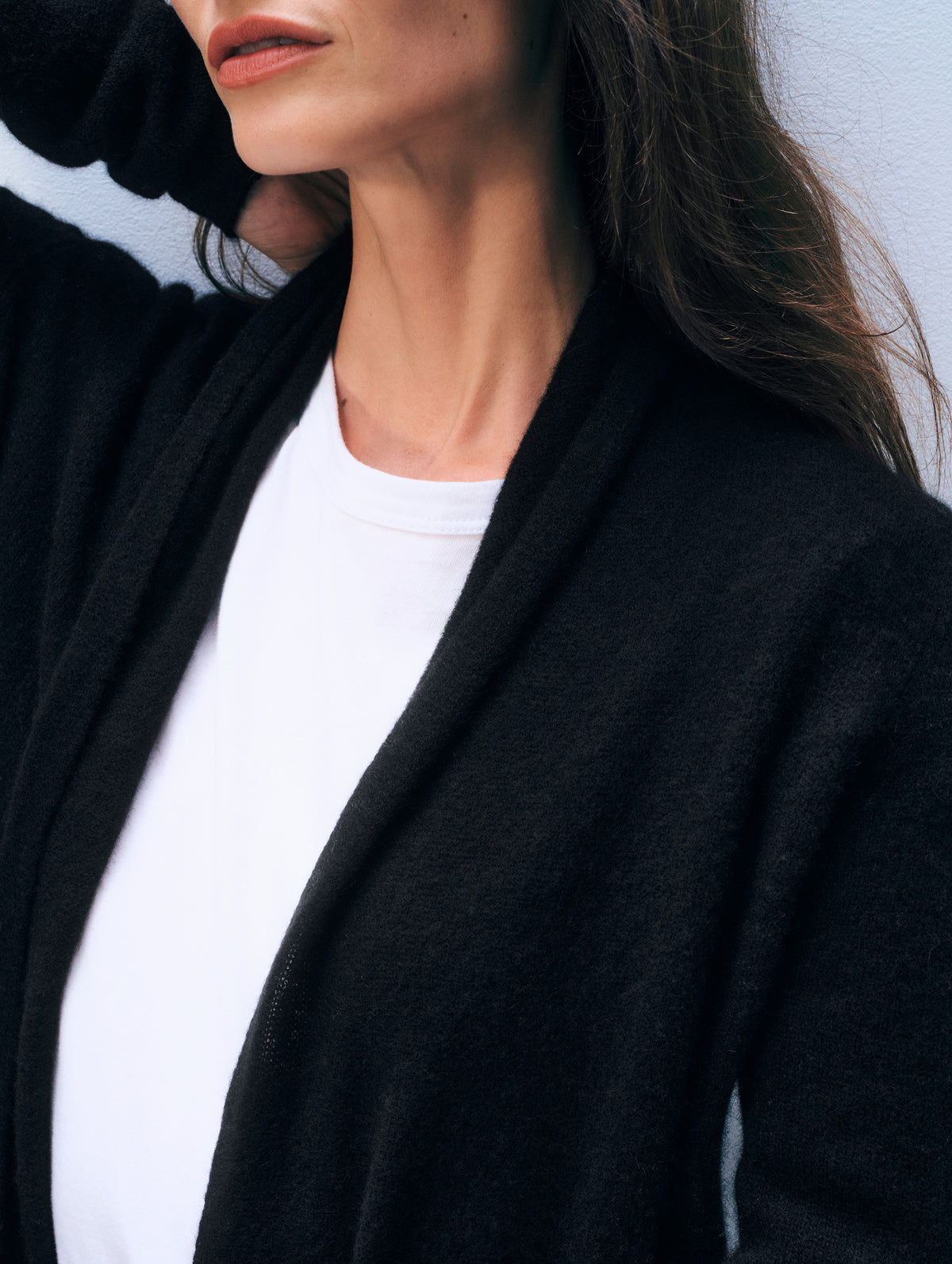 A close-up of a woman wearing a black open-front cardigan over a white crew-neck t-shirt, with her hair and lower face visible against a light wall.