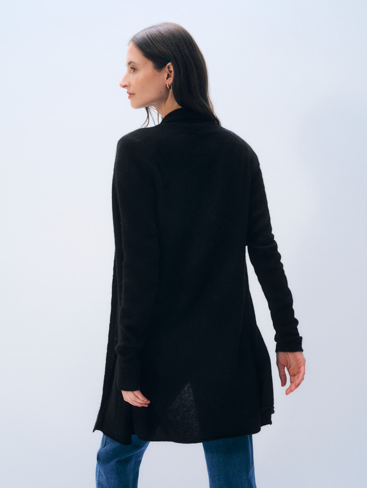 A woman wearing a black long-sleeve turtleneck cardigan, shown from a side-back angle against a light gray studio background.