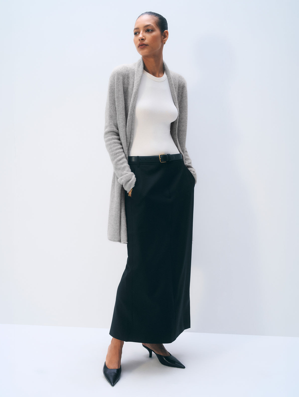 A woman models a stylish outfit consisting of a light gray knit cardigan, a white ribbed top, a long black skirt, and black pointed-toe heels, set against a clean white studio background.