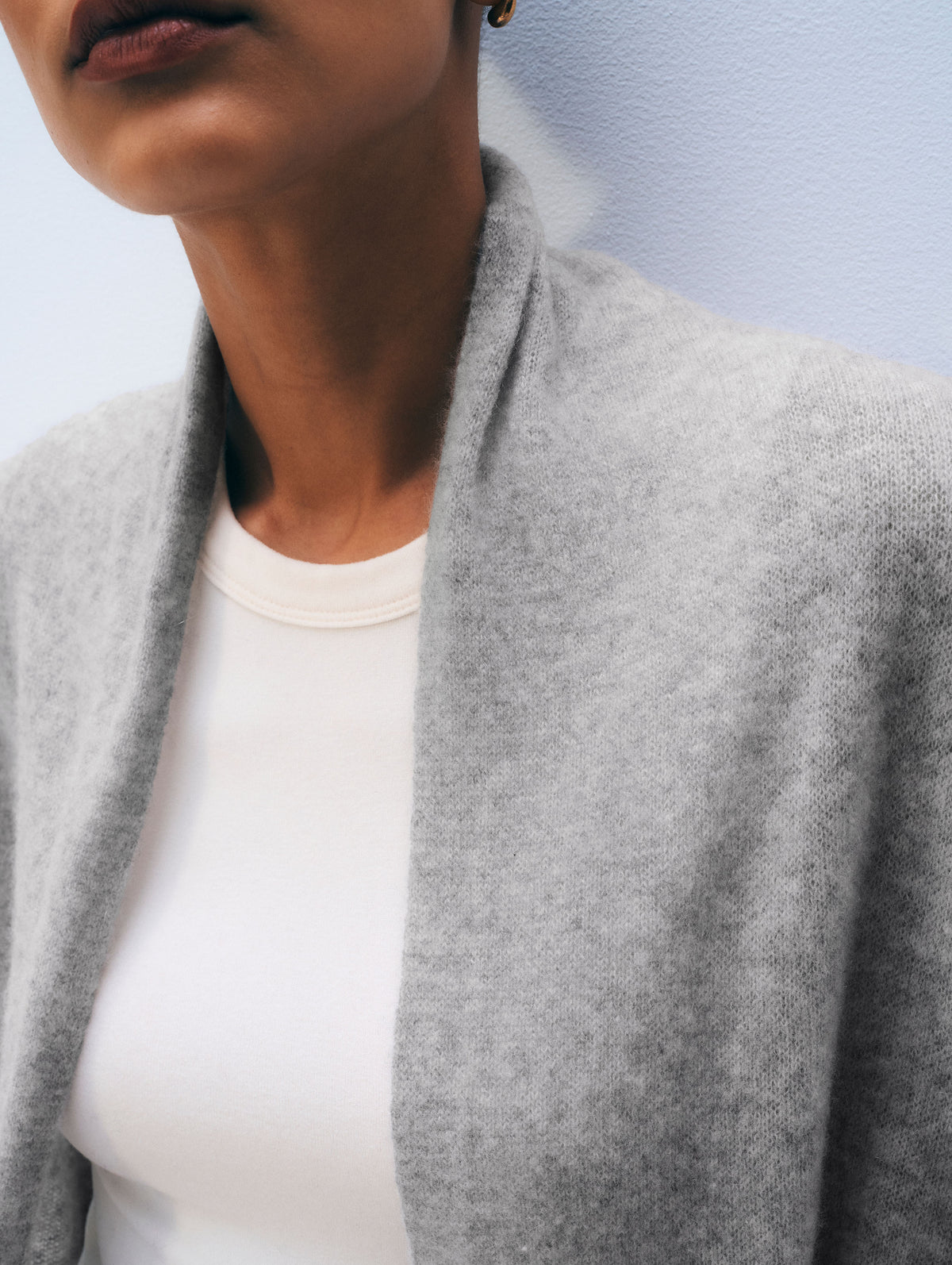 Close-up of a person wearing a light gray knit cardigan or wrap over a white top, against a pale blue wall.
