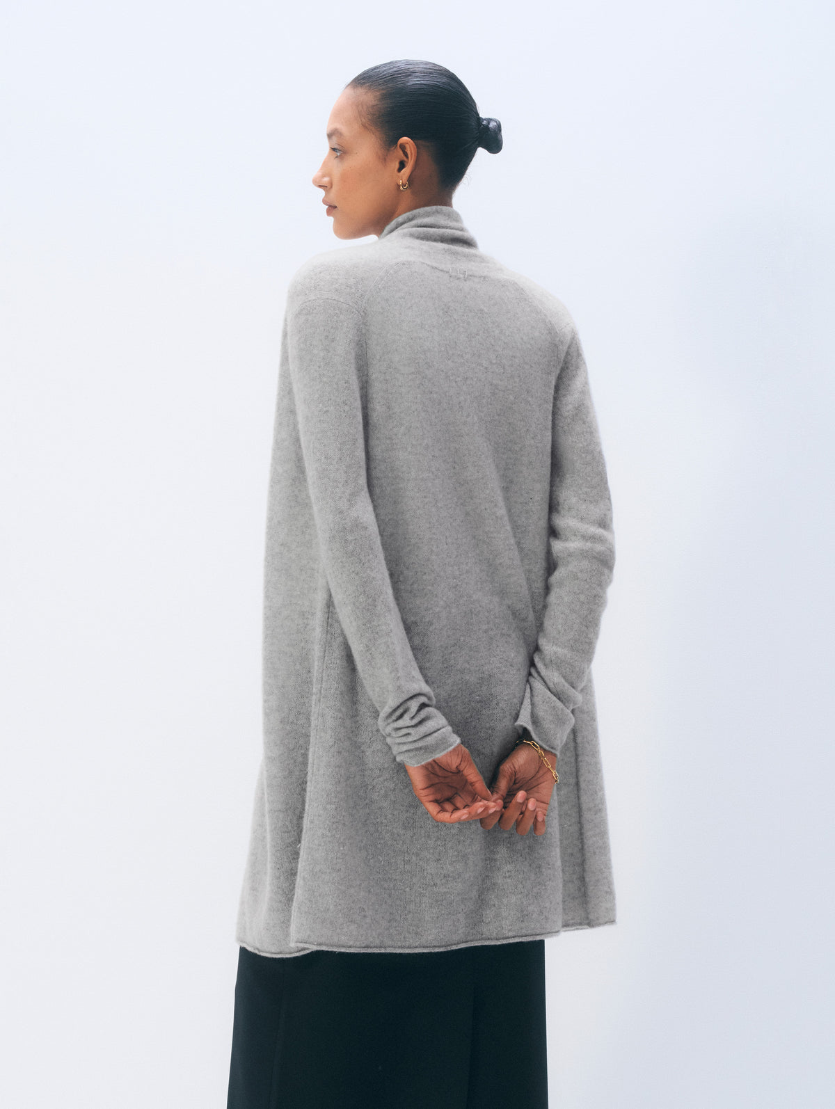 A model shown from the back wearing a light gray knit cowl-neck cardigan with long sleeves and a draped silhouette, paired with black wide-leg trousers, against a clean white studio background.