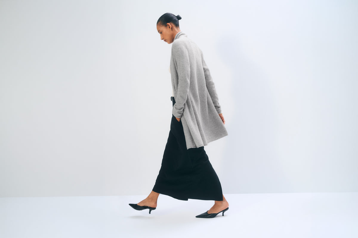 A woman walking in profile wearing a light gray long-sleeve cardigan over a long black skirt, paired with black pointed-toe flats, against a clean white studio background.