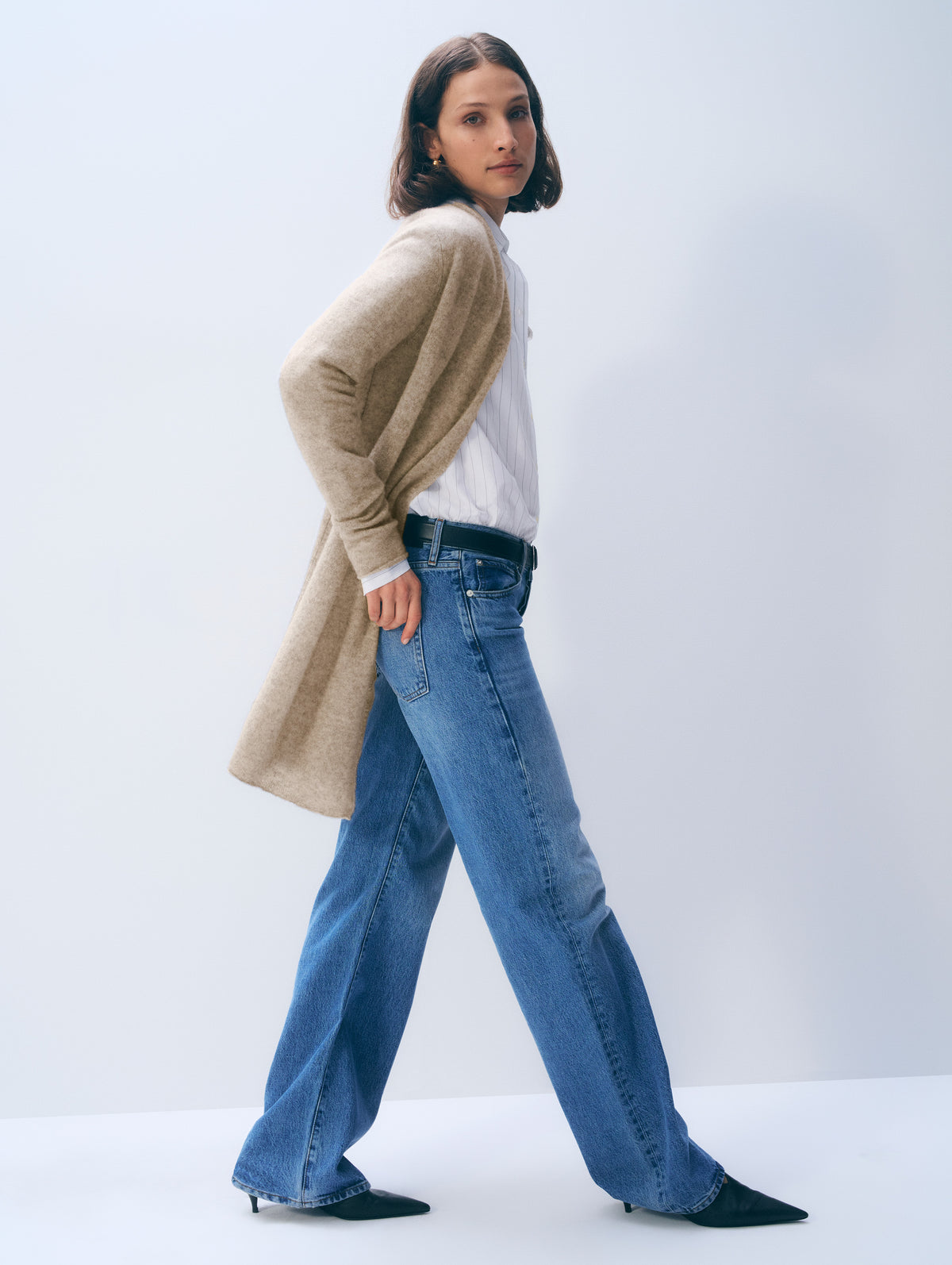 A woman modeling a beige knit cardigan over a white top and wide-leg blue jeans, standing in a studio with a light gray background.
