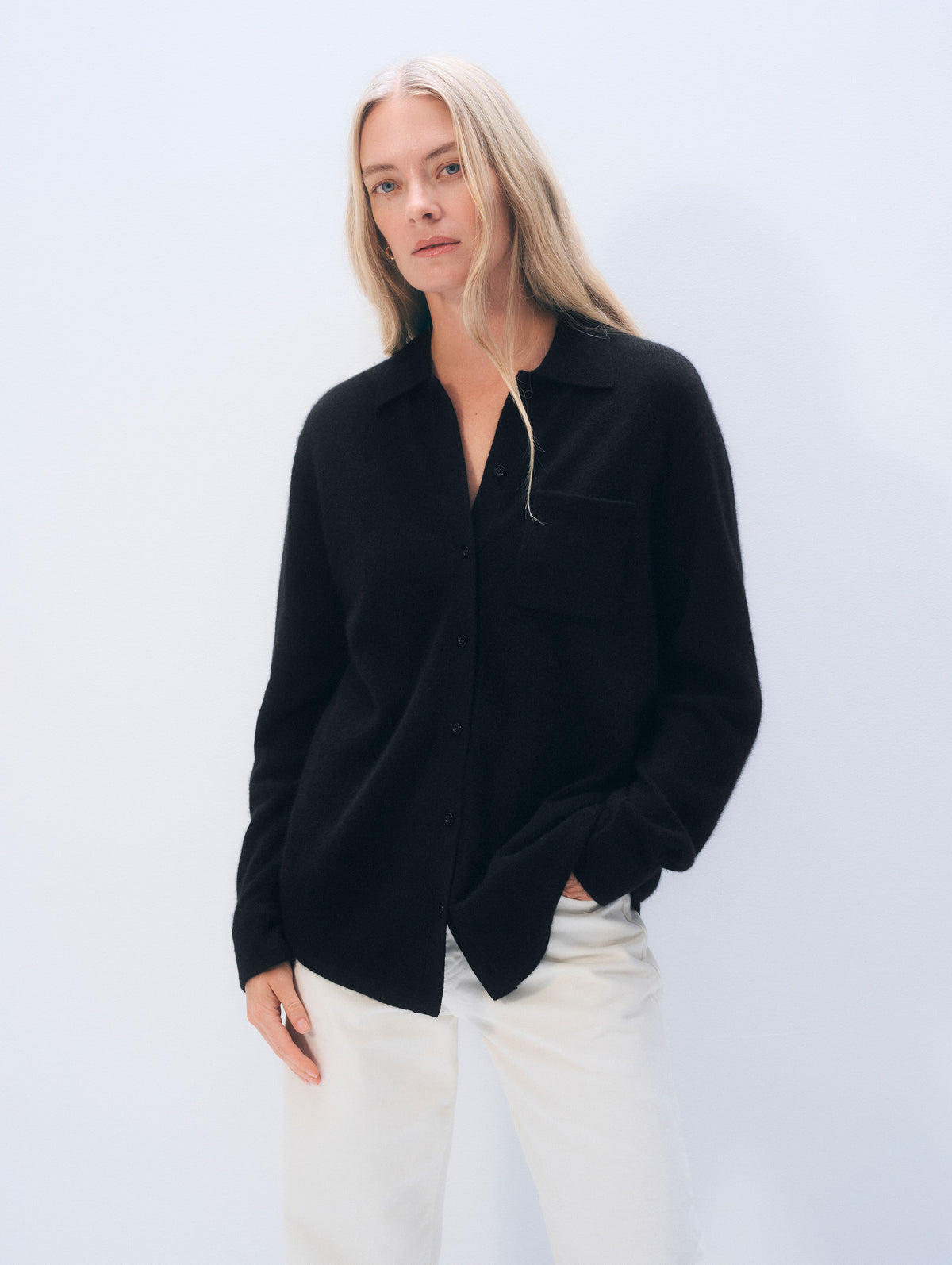 A woman modeling a black button-up shirt with a relaxed fit and white pants against a light blue studio background.