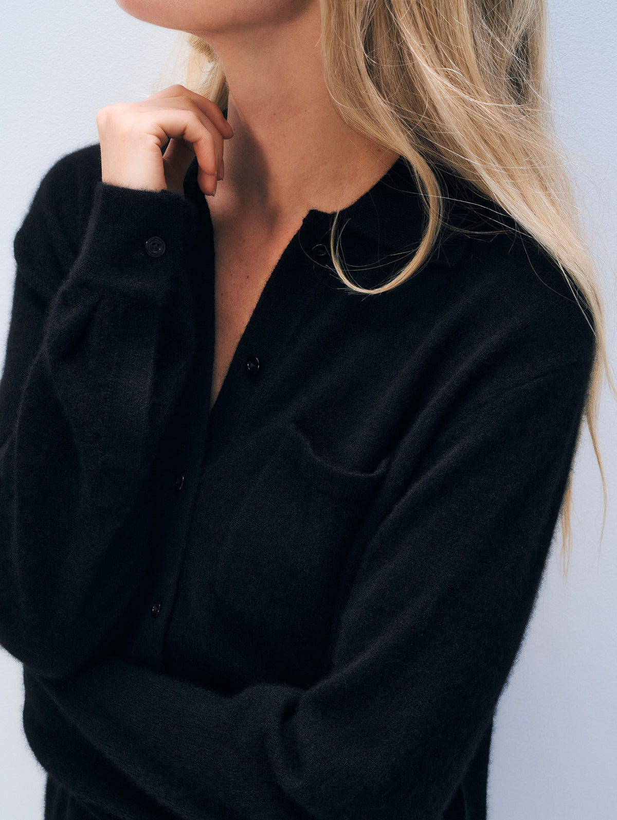 A close-up of a woman wearing a black long-sleeve knit button-front top with a chest pocket, shown against a light gray background.