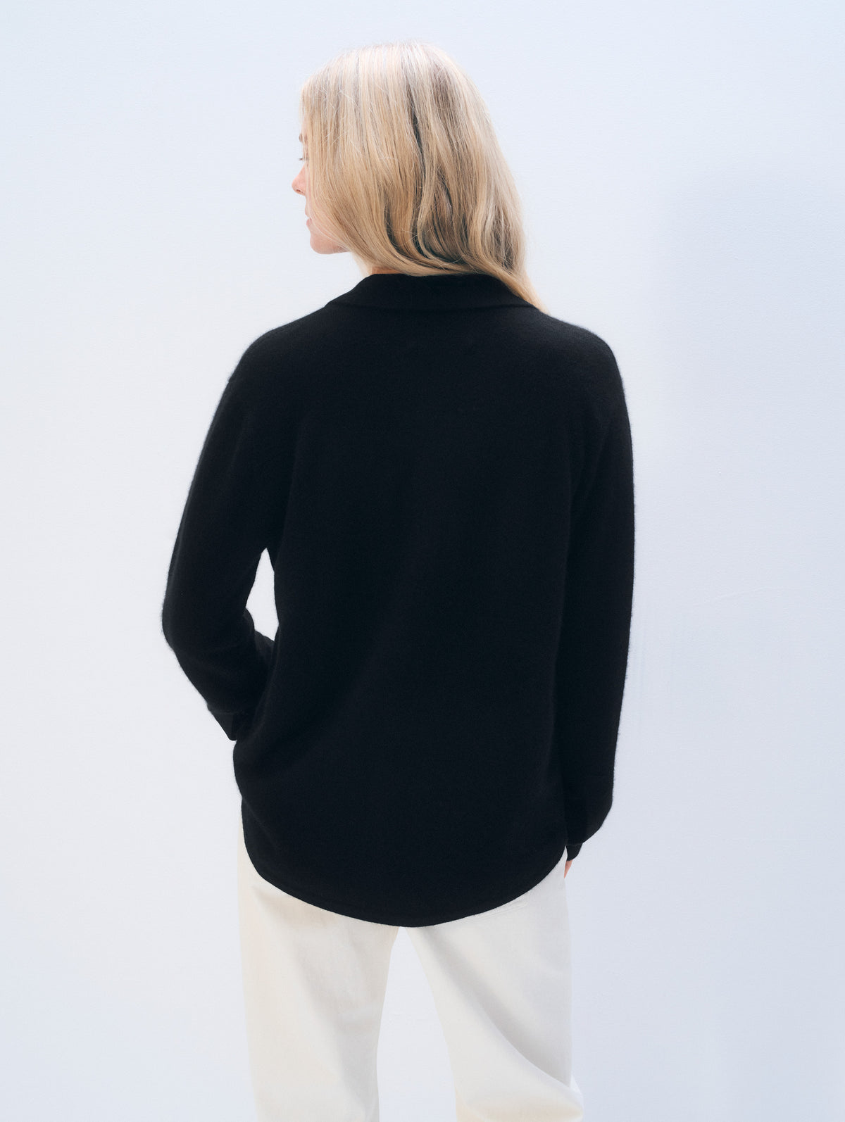 A woman with blonde hair is shown from the back wearing a black long-sleeve knit top and cream-colored pants against a light gray studio background.