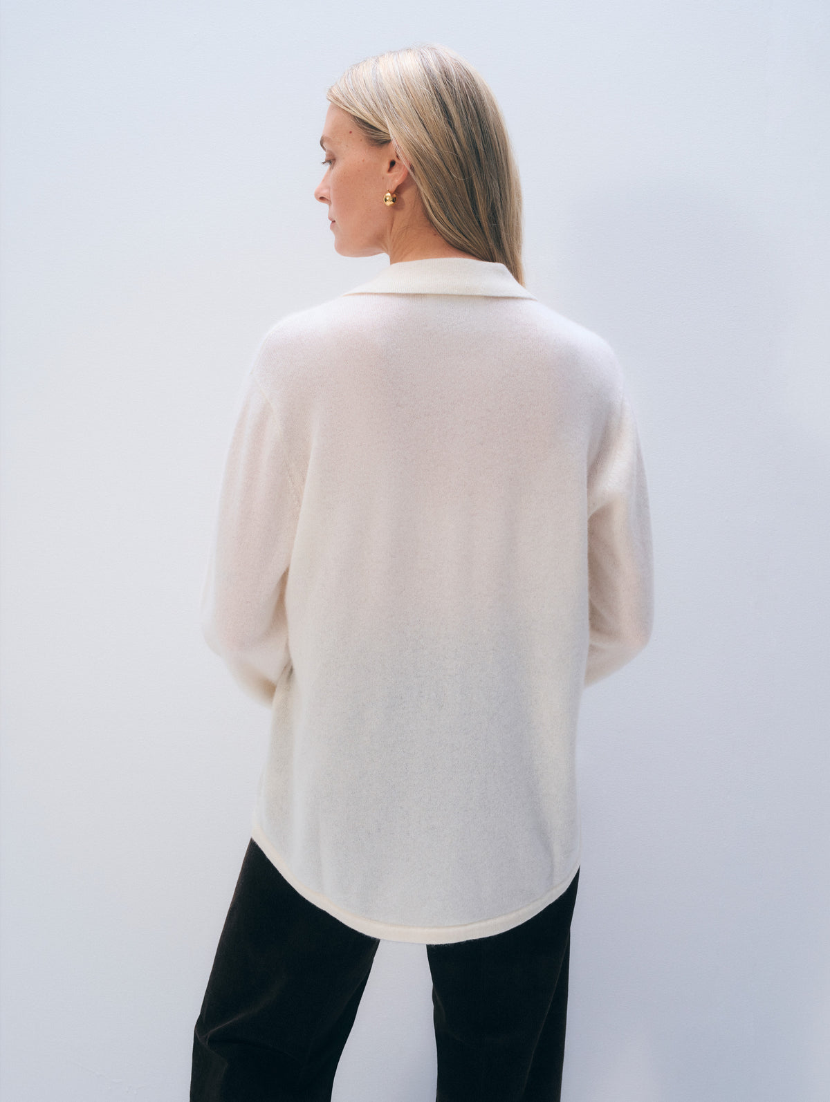 A woman with blonde hair is shown from the back wearing a cream-colored knit long-sleeve top and black pants, standing against a light gray studio background.