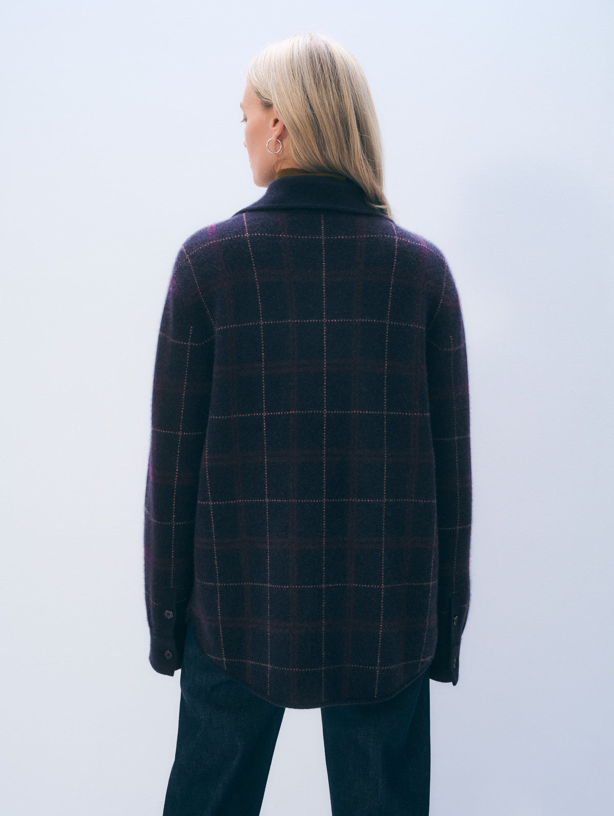 A woman viewed from behind wearing a dark navy and purple plaid wool-blend jacket with a high collar, paired with dark jeans, against a light gray studio background.