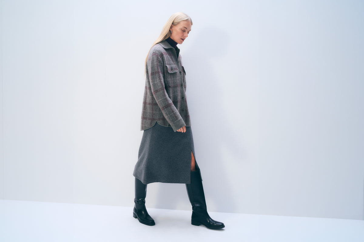 A woman modeling a long-sleeve gray plaid overshirt over a black turtleneck, paired with a dark gray midi skirt and black knee-high boots, set against a plain light gray studio background.