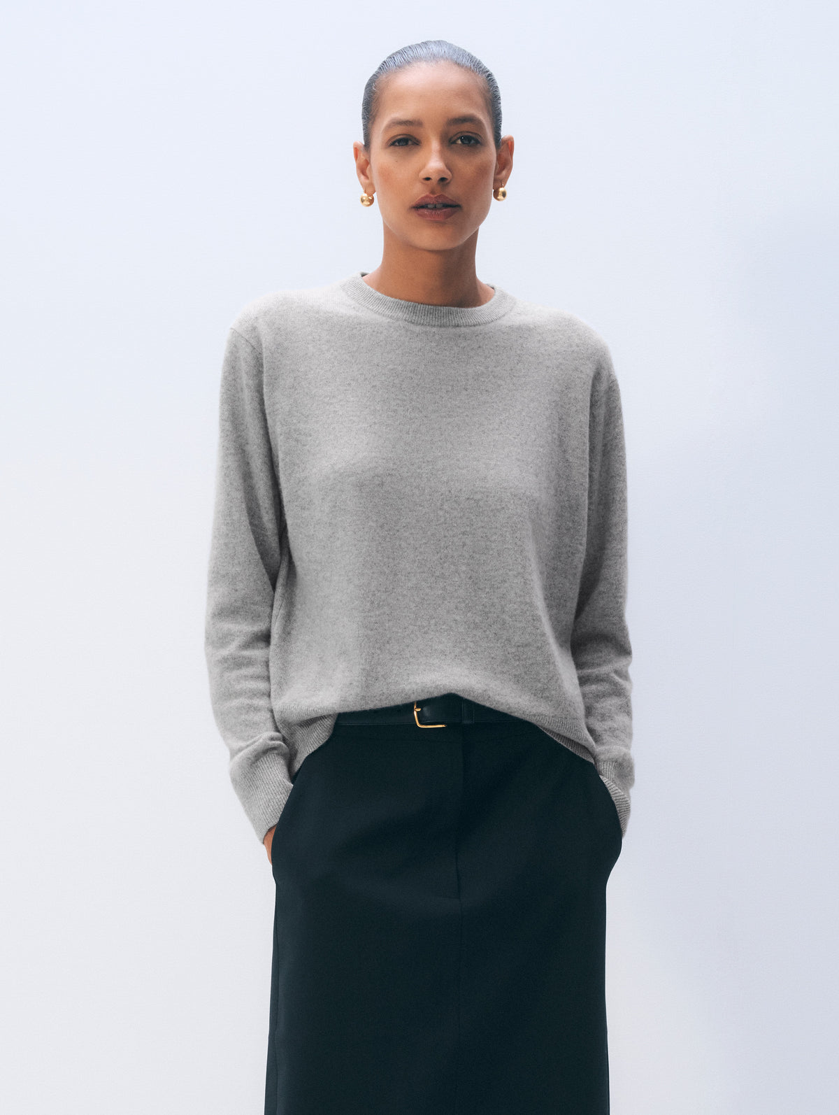 A woman modeling a light gray crewneck knit sweater paired with black trousers and a thin gold-tone belt, set against a clean, light gray studio background.