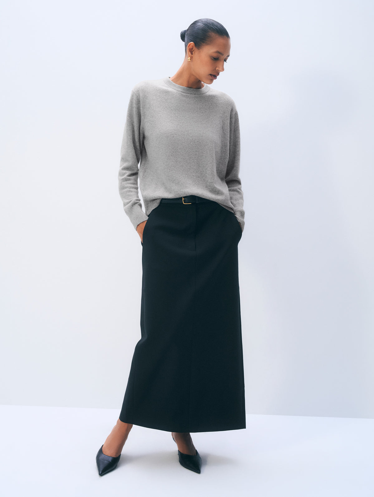 A woman modeling a light gray knit sweater and a long black skirt, standing against a clean white studio background.
