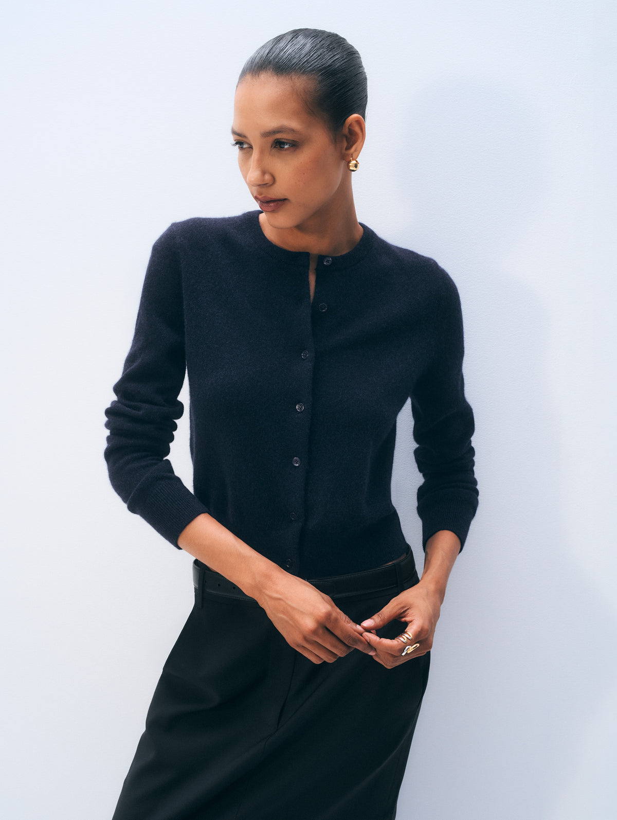 A woman modeling a black long-sleeve knit cardigan with a buttoned front and a matching black skirt against a light gray studio background.