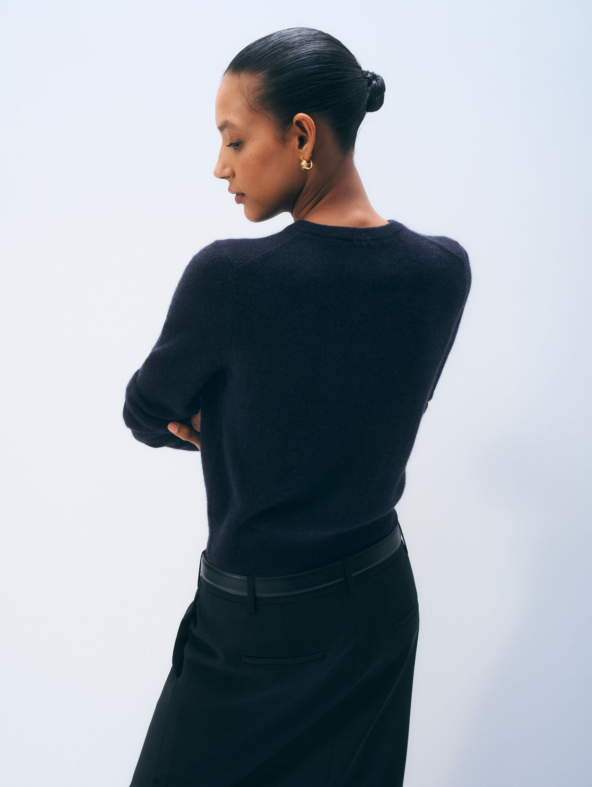 A model shown from the back wearing a dark navy knit sweater and a black skirt, set against a clean, light gray studio background.