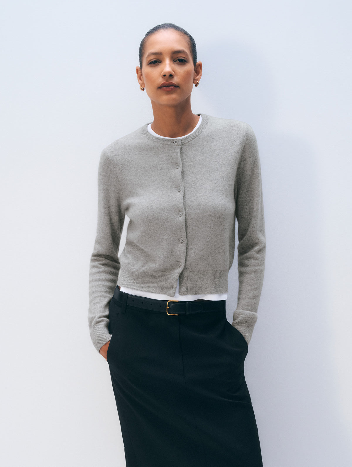 A woman modeling a light gray cropped button-front cardigan over a white top and black skirt, standing against a plain light gray studio background.