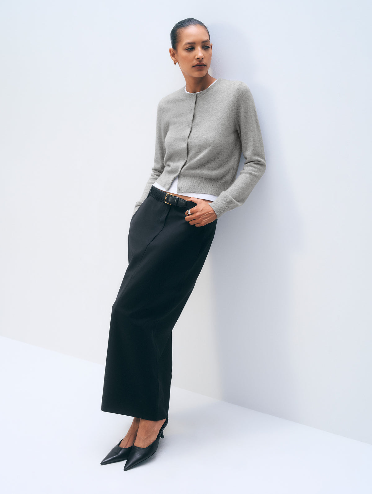 A model wearing a light gray zip-front cardigan, black wide-leg trousers, and black pointed-toe heels, leaning against a white studio wall.