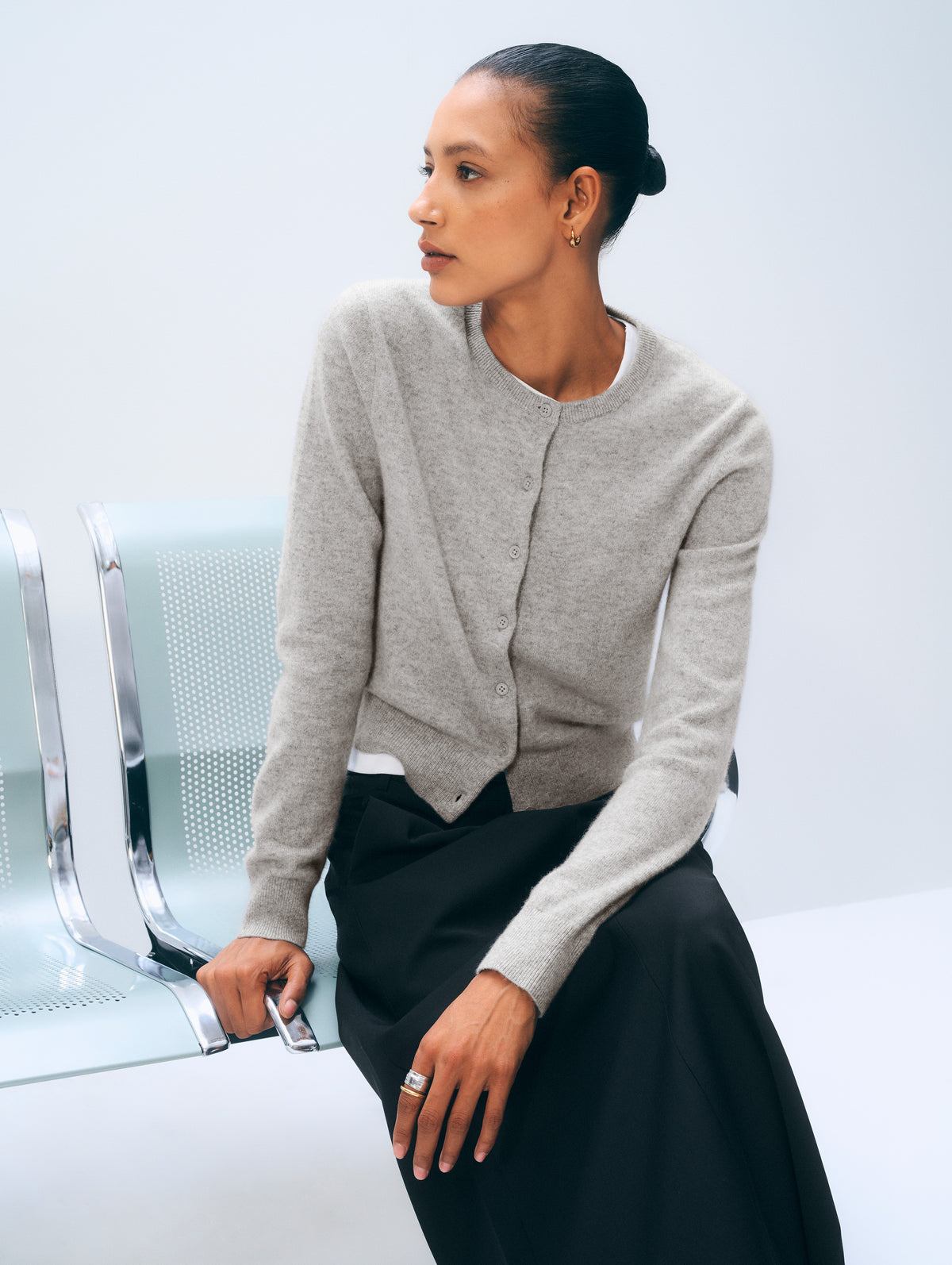A woman wearing a light gray knit cardigan and a black skirt sits on a modern light blue bench against a pale studio background.