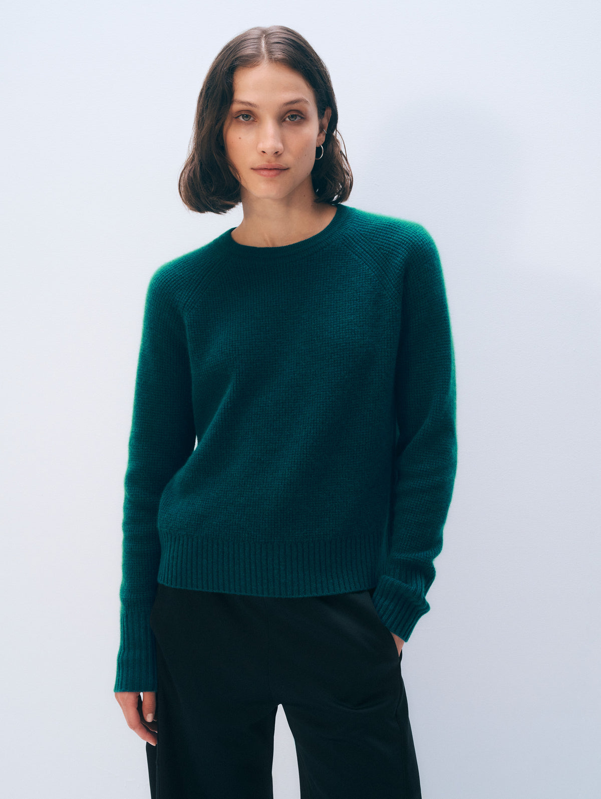 A woman wearing a dark teal-green knit sweater and black trousers, standing against a light gray studio background.