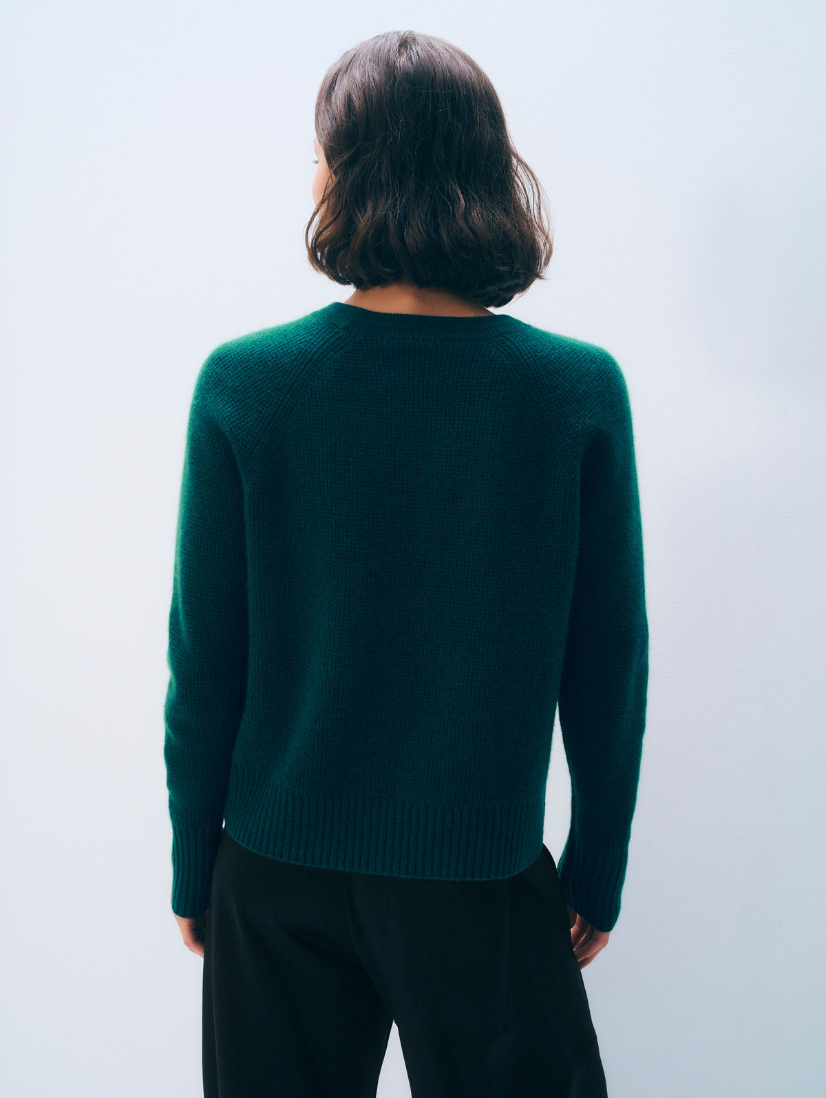 A person shown from the back wearing a dark teal-green knit sweater and black pants against a plain light gray studio background.