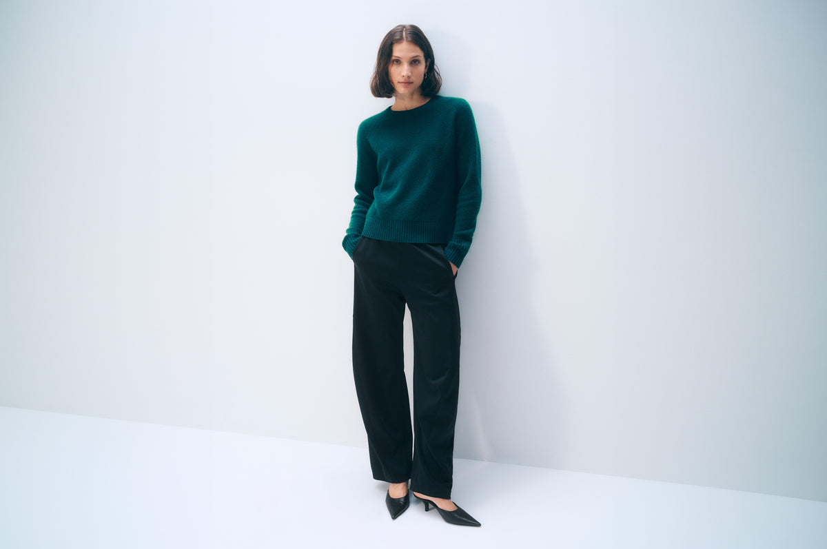 A woman stands against a plain white studio wall wearing a dark green long-sleeve knit sweater, black wide-leg trousers, and black pointed shoes.