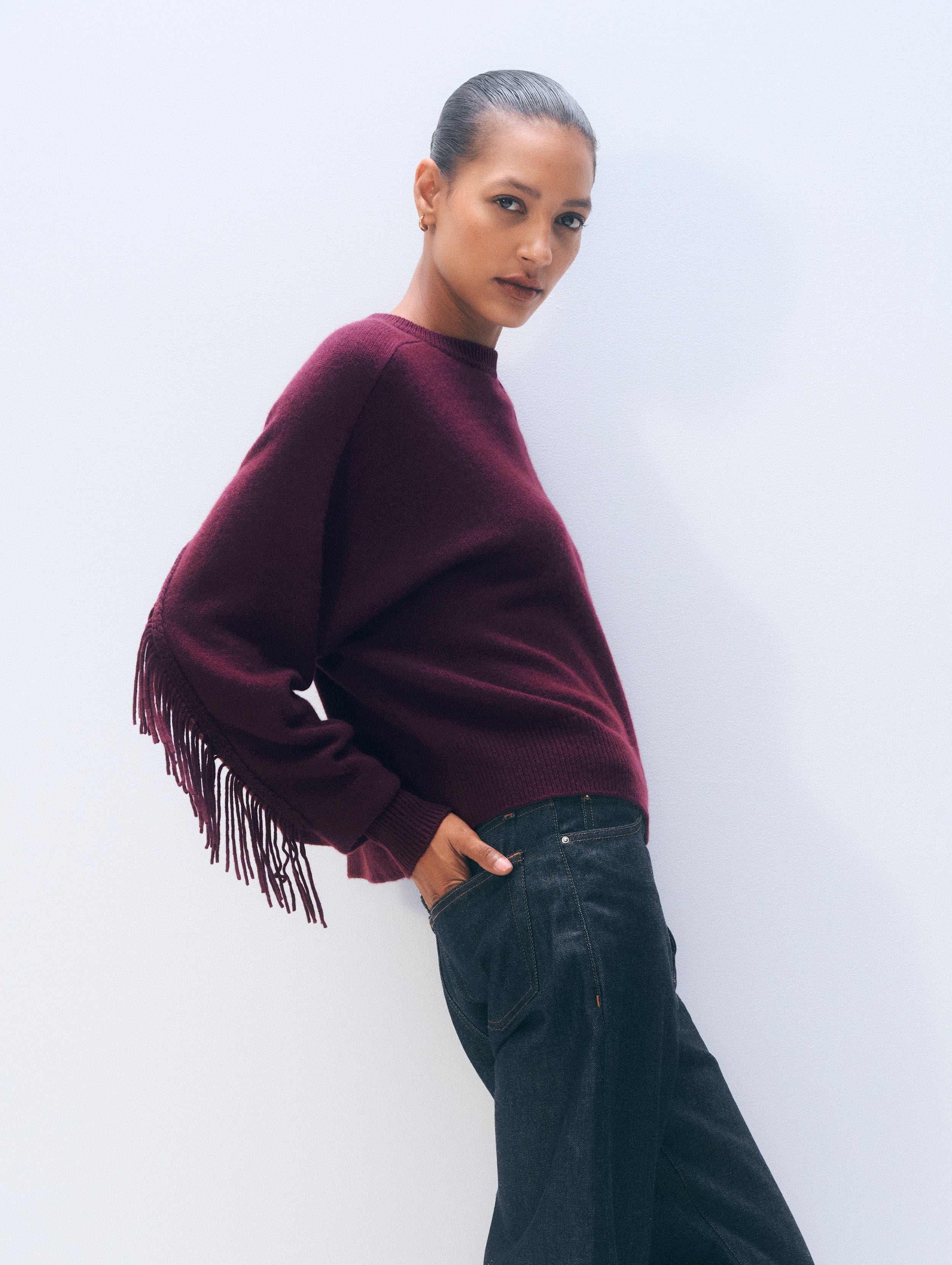 A woman modeling a deep burgundy knit sweater with fringe detailing on the sleeve, paired with dark wide-leg jeans against a clean white studio background.