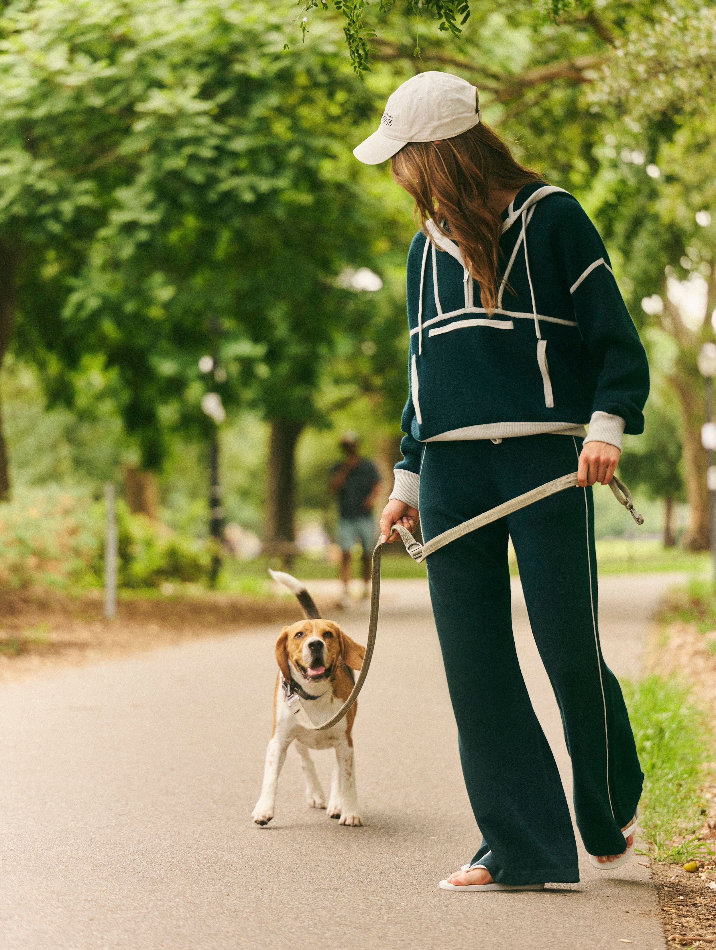 A woman wearing a dark teal two-piece tracksuit with white stripes and a white baseball cap walks a beagle on a paved path in a park.