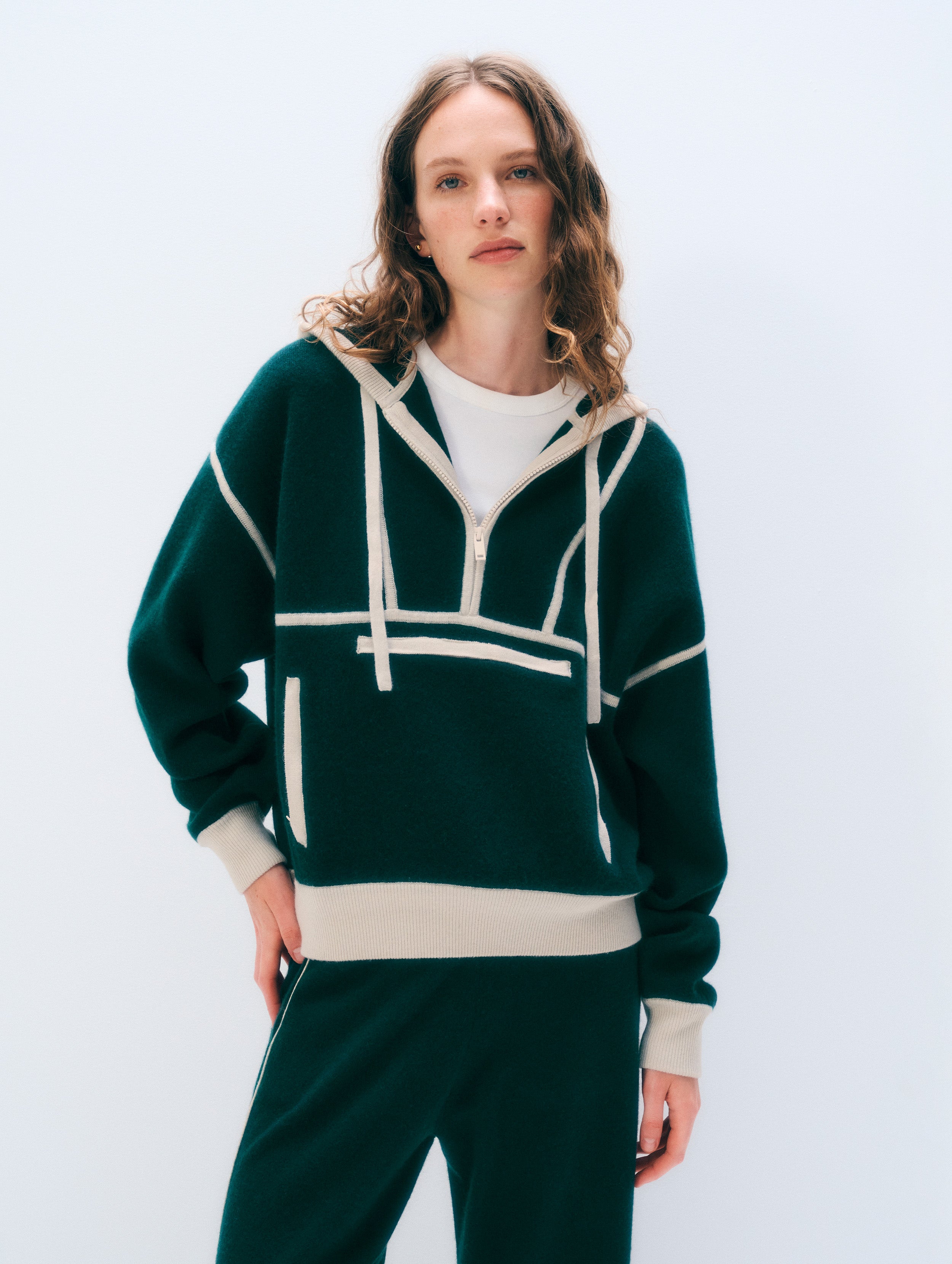 A woman modeling a dark green half-zip hoodie with cream-colored geometric stripe details, a cream hem, and a white inner shirt, paired with matching dark green sweatpants against a light gray studio background.