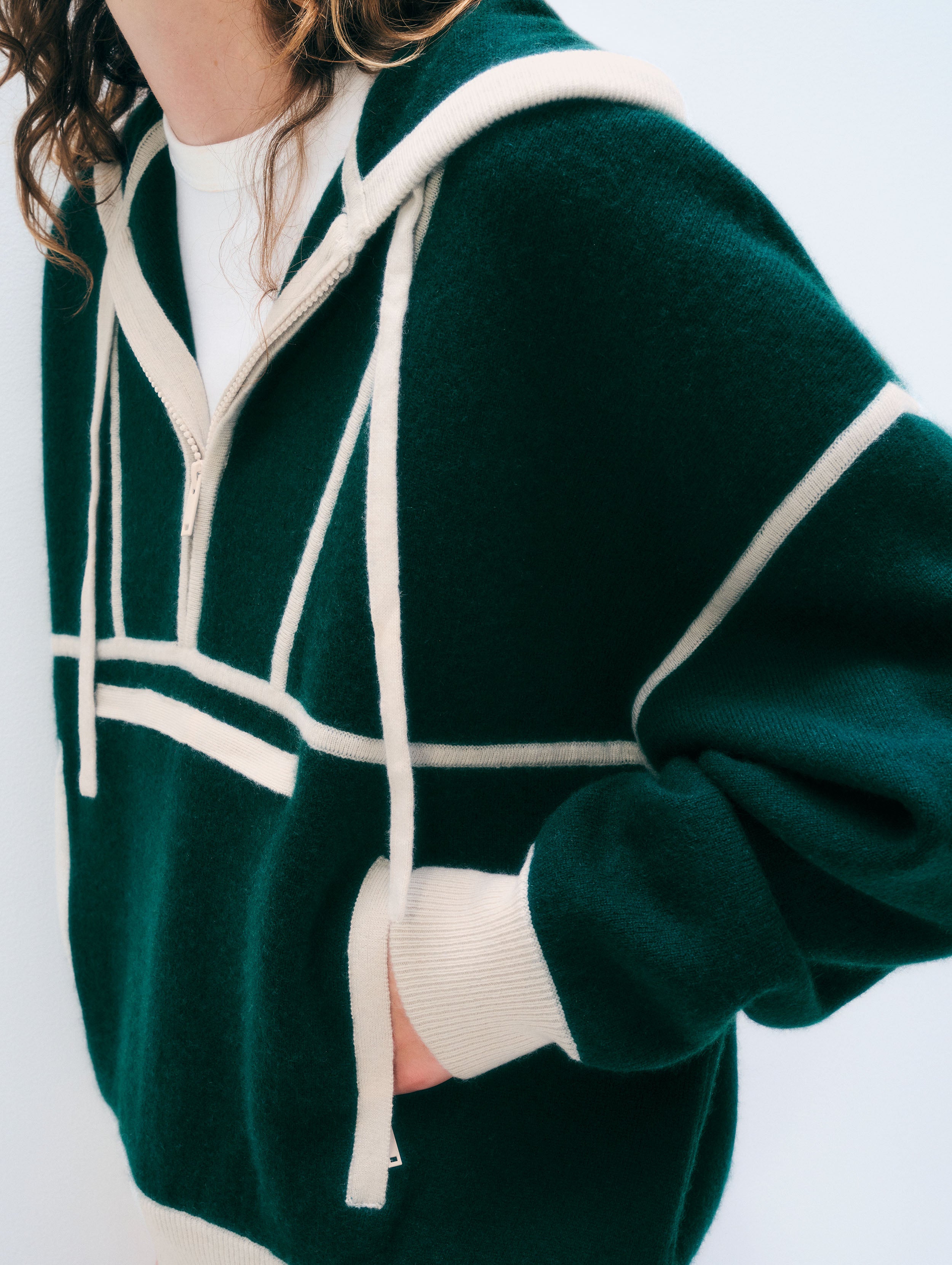 A close-up of a person wearing a dark green knit zip-up hoodie with contrasting cream-colored trim, drawstrings, and ribbed cuffs, layered over a white top.
