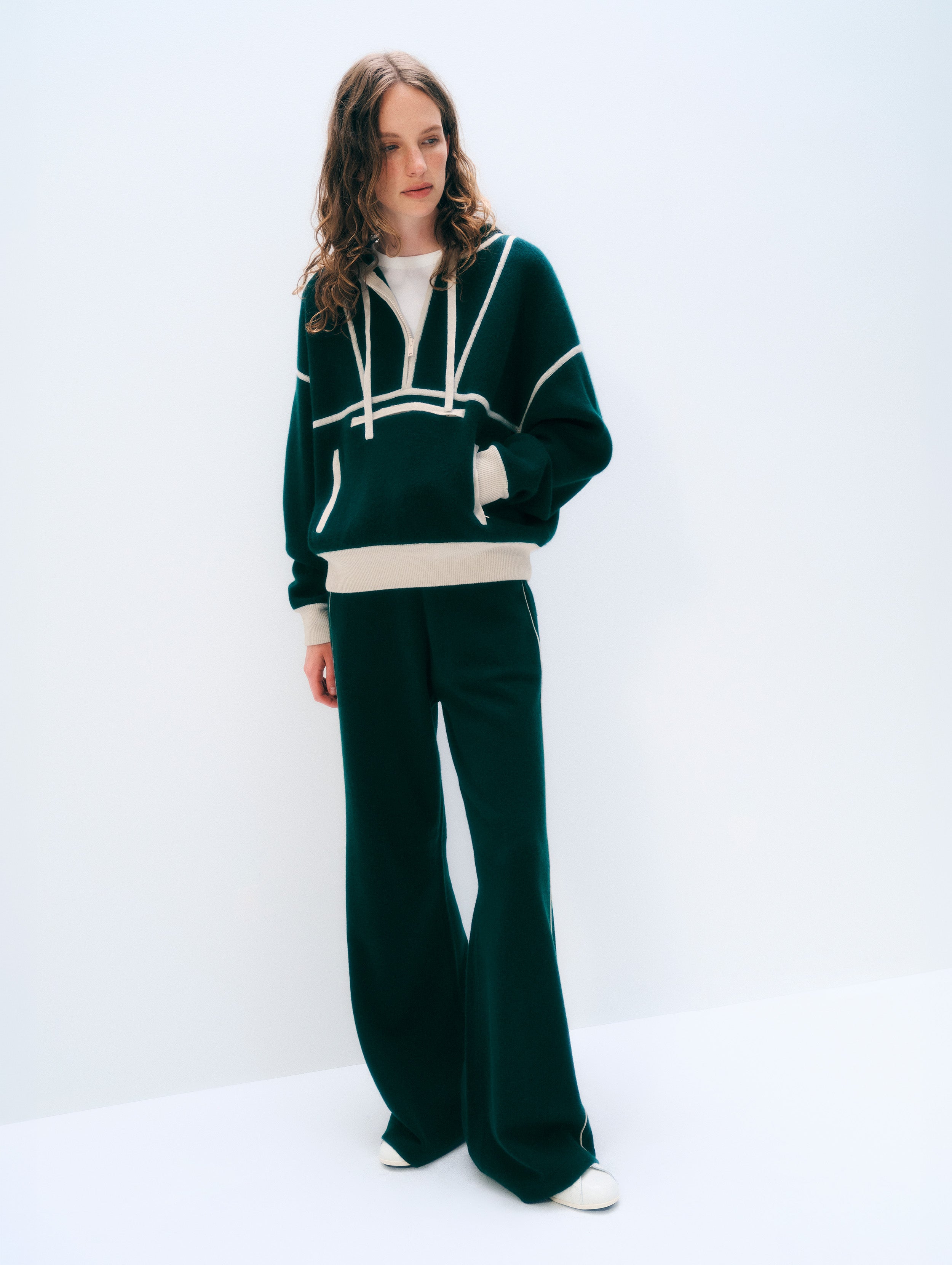 A woman modeling a dark green knit tracksuit with white contrast stitching and white drawstrings, featuring a cropped hoodie top and matching wide-leg pants, set against a plain white studio background.