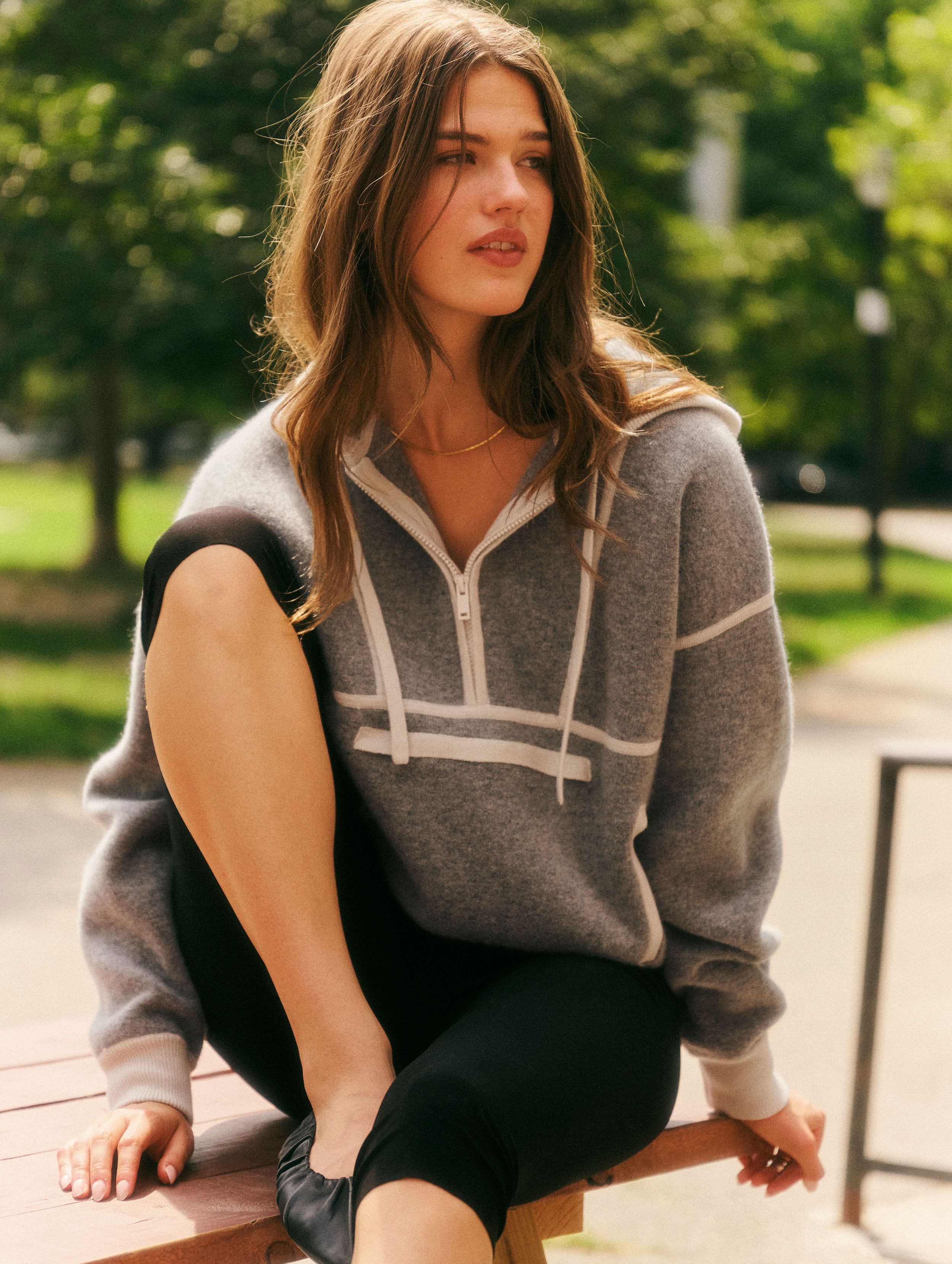 A woman sitting outdoors on a bench wearing a gray half-zip hoodie with white stripe details, black leggings, and black shoes, with a blurred park background.