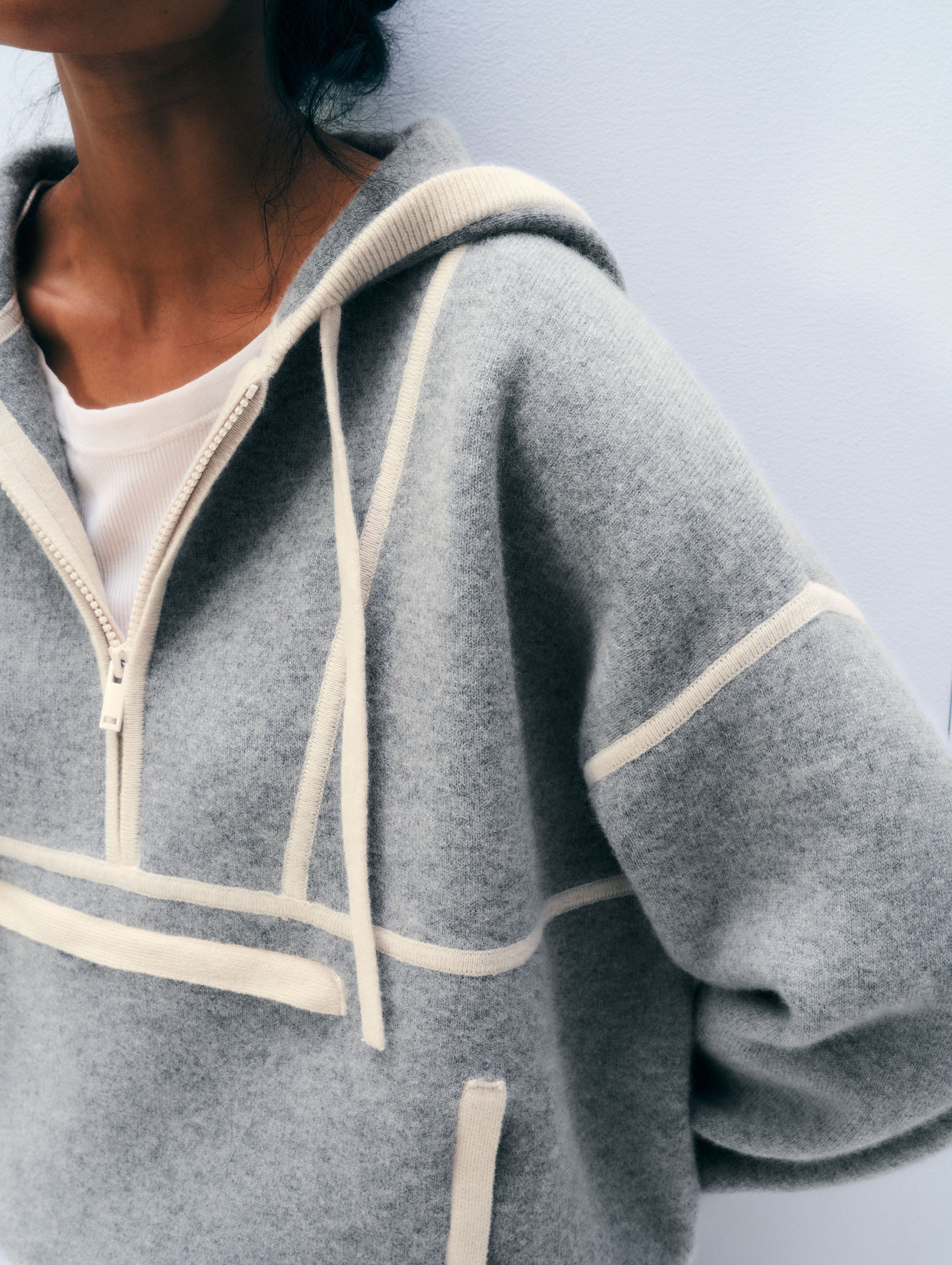 Close-up of a person wearing a heather gray zip-up hoodie with cream-colored contrast stitching and drawstrings over a white t-shirt, set against a plain light gray wall.