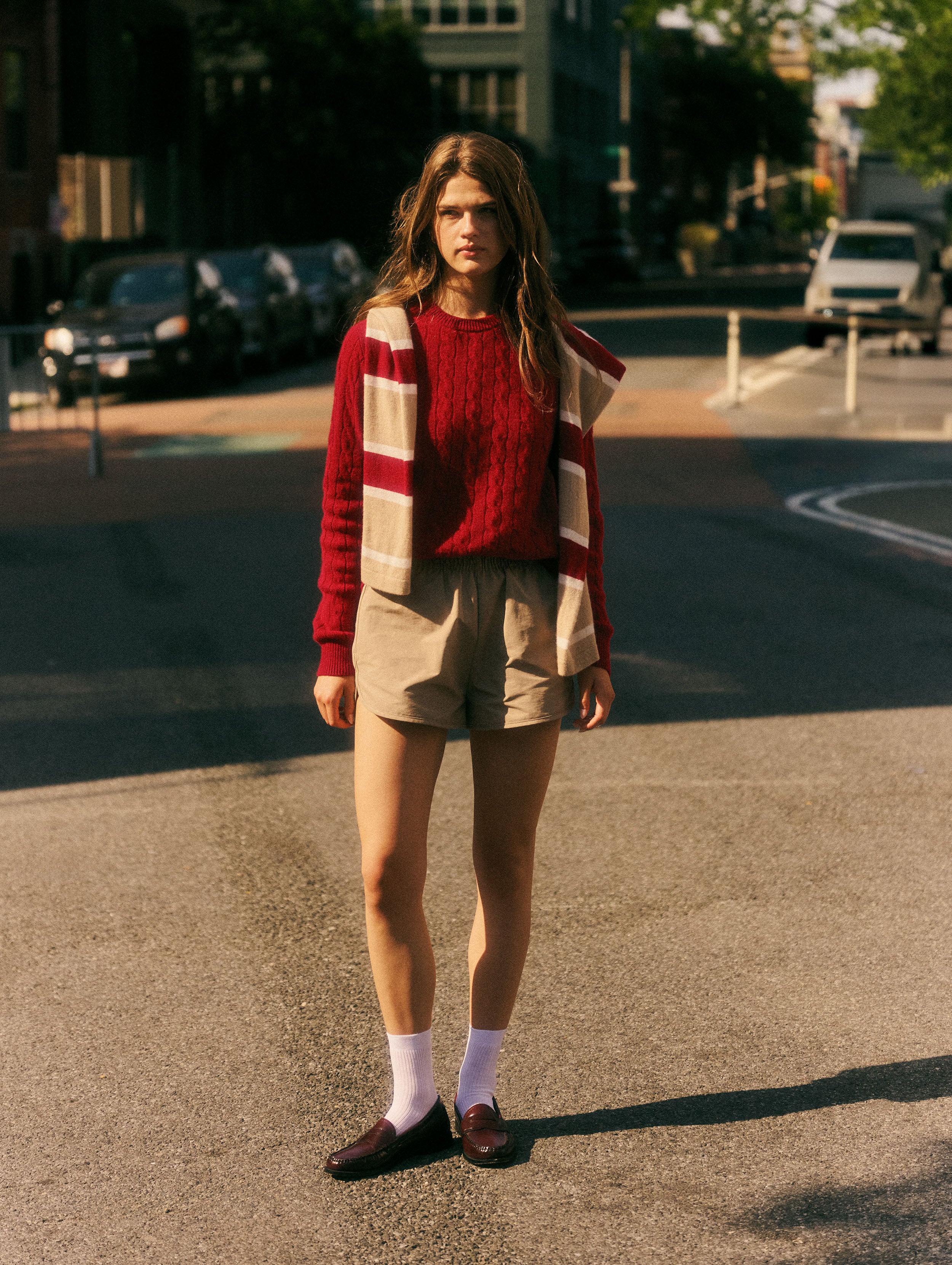 A woman stands on an urban street wearing a red cable-knit sweater, a red and beige striped cardigan, beige shorts, white socks, and dark loafers.