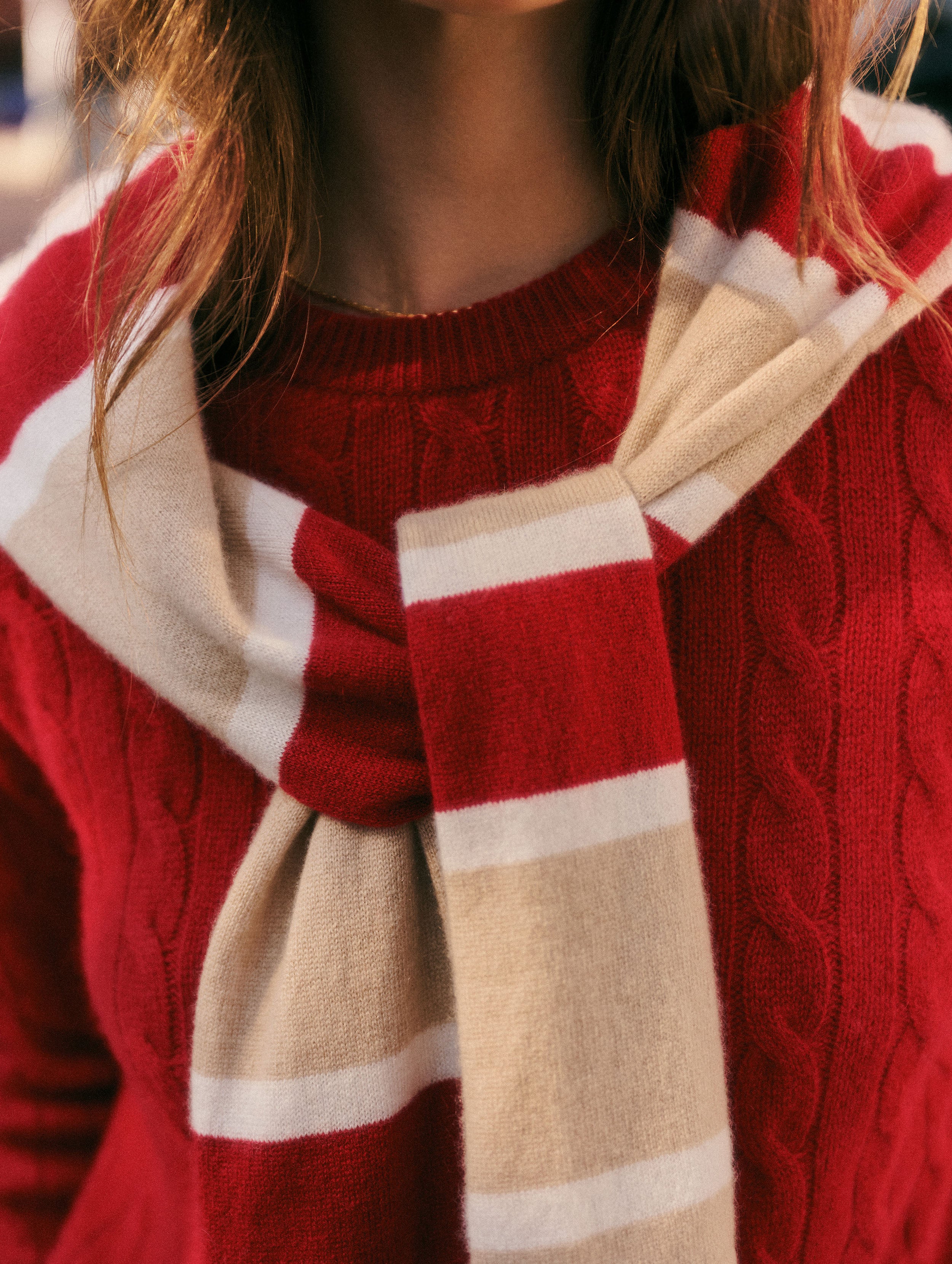 A close-up of a person wearing a red cable-knit sweater and a cream and white striped knit scarf.