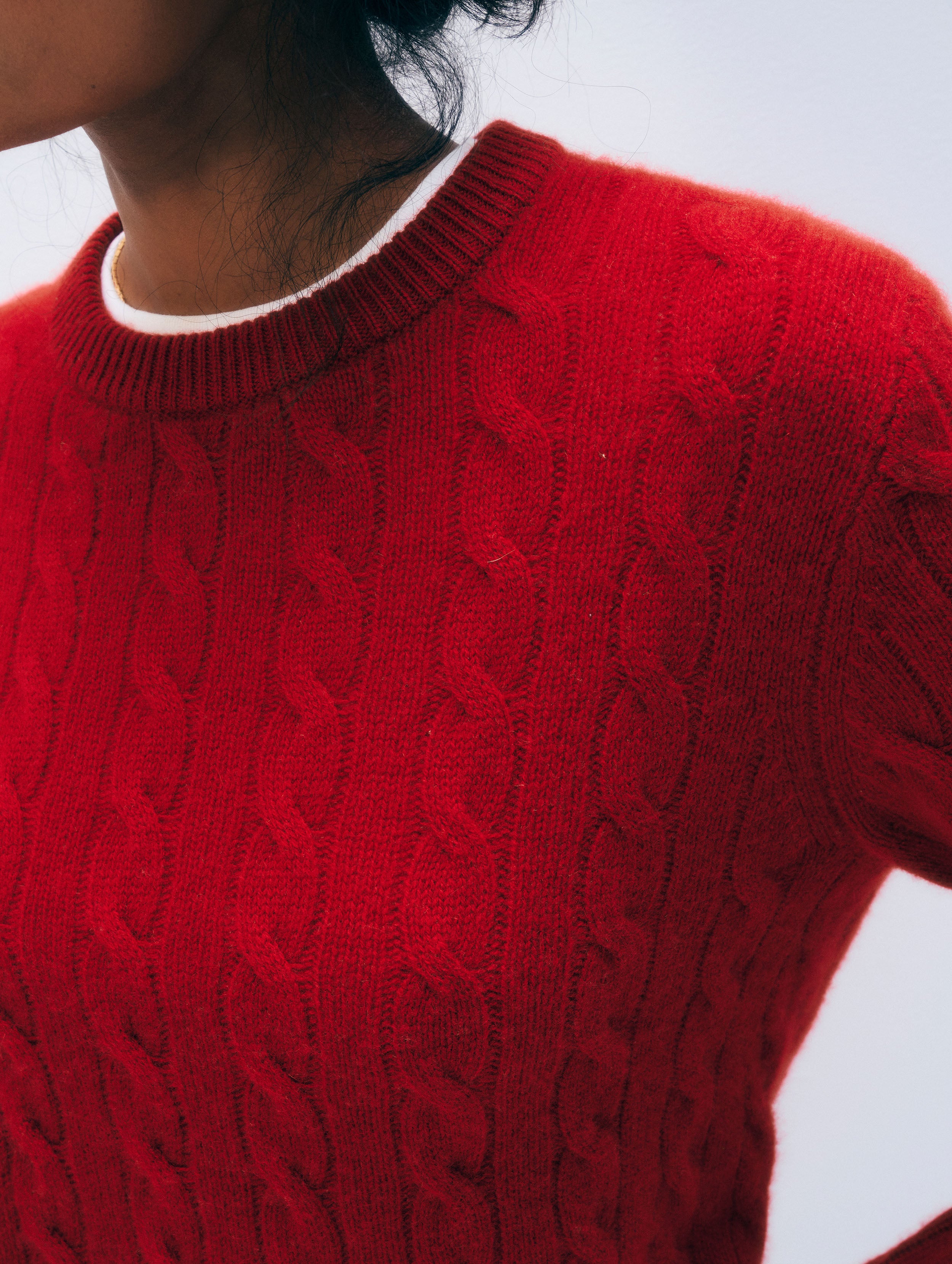 A close-up of a person wearing a red cable-knit sweater with a white crew neck collar visible underneath, against a light gray background.
