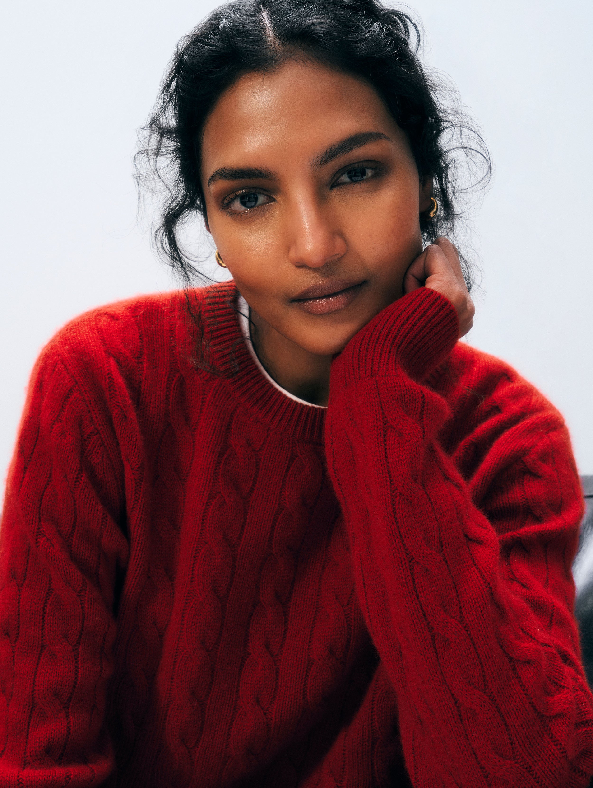 A close-up portrait of a woman wearing a vibrant red cable-knit crewneck sweater, resting her chin on her hand against a plain light gray background.