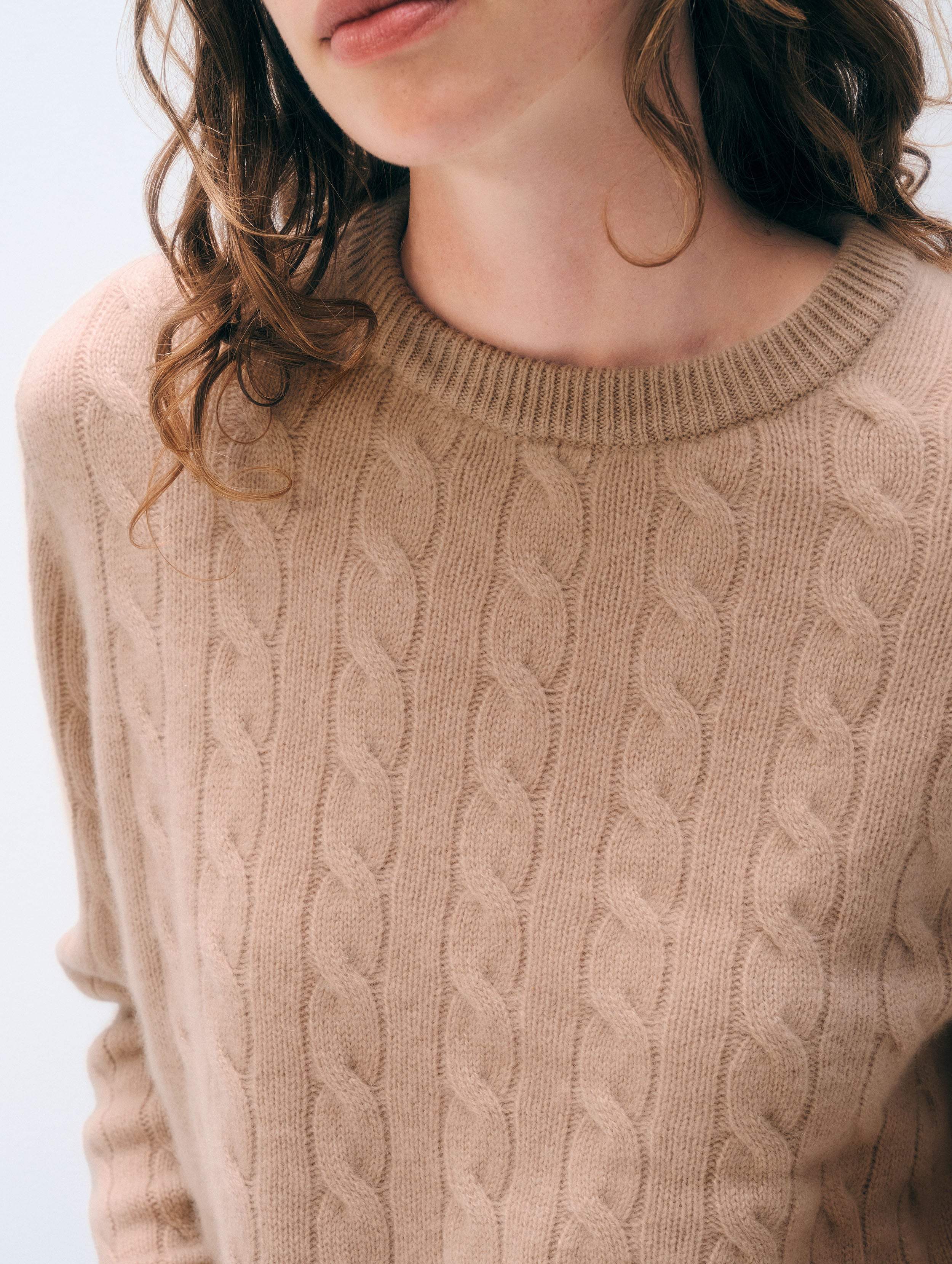 Close-up of a woman wearing a beige cable-knit crewneck sweater, showing the detailed knit texture and her hair against a light background.