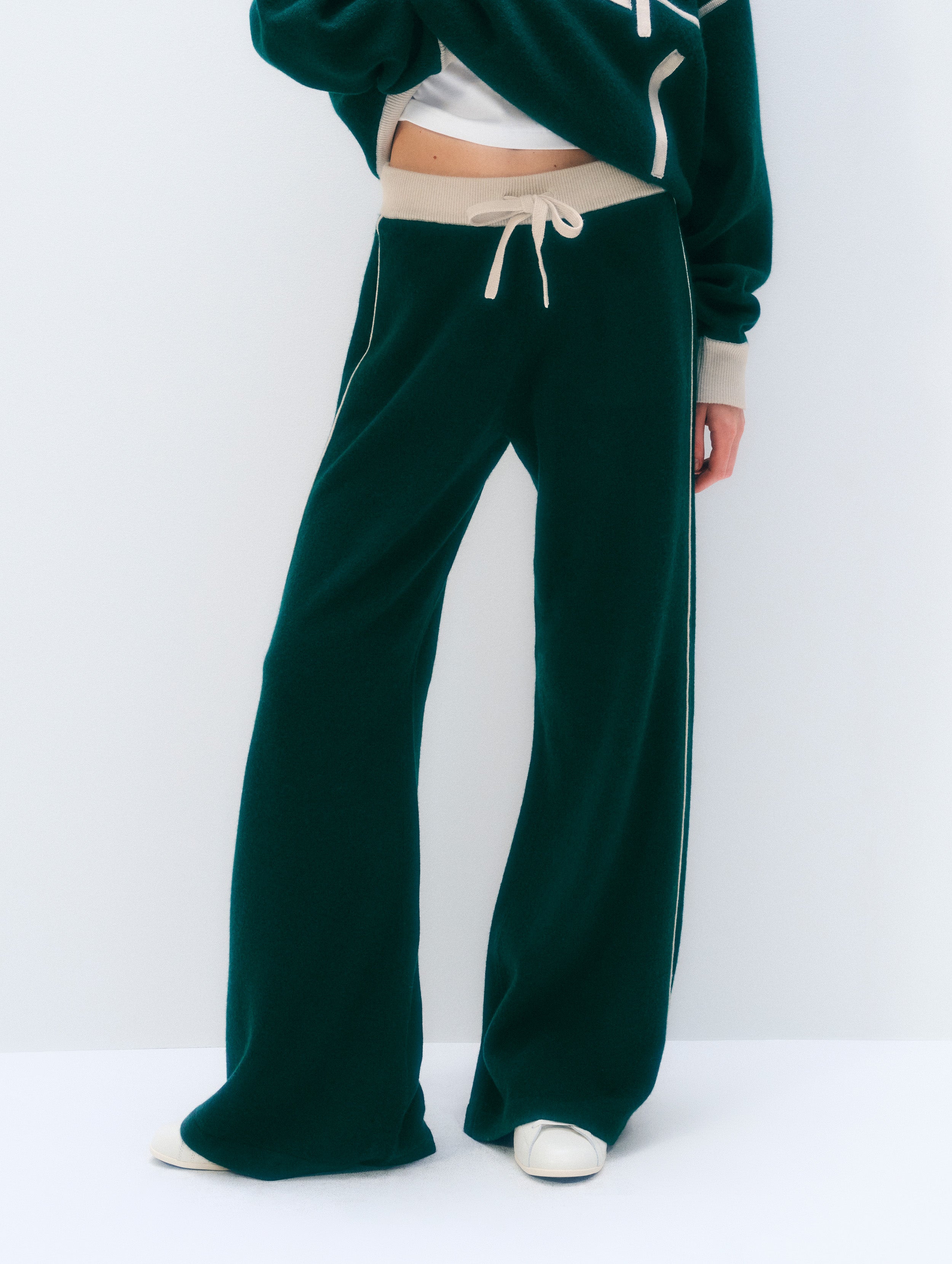 A person wearing dark green wide-leg sweatpants with a beige drawstring waistband and a matching dark green cropped sweatshirt, shown against a plain light gray studio background.