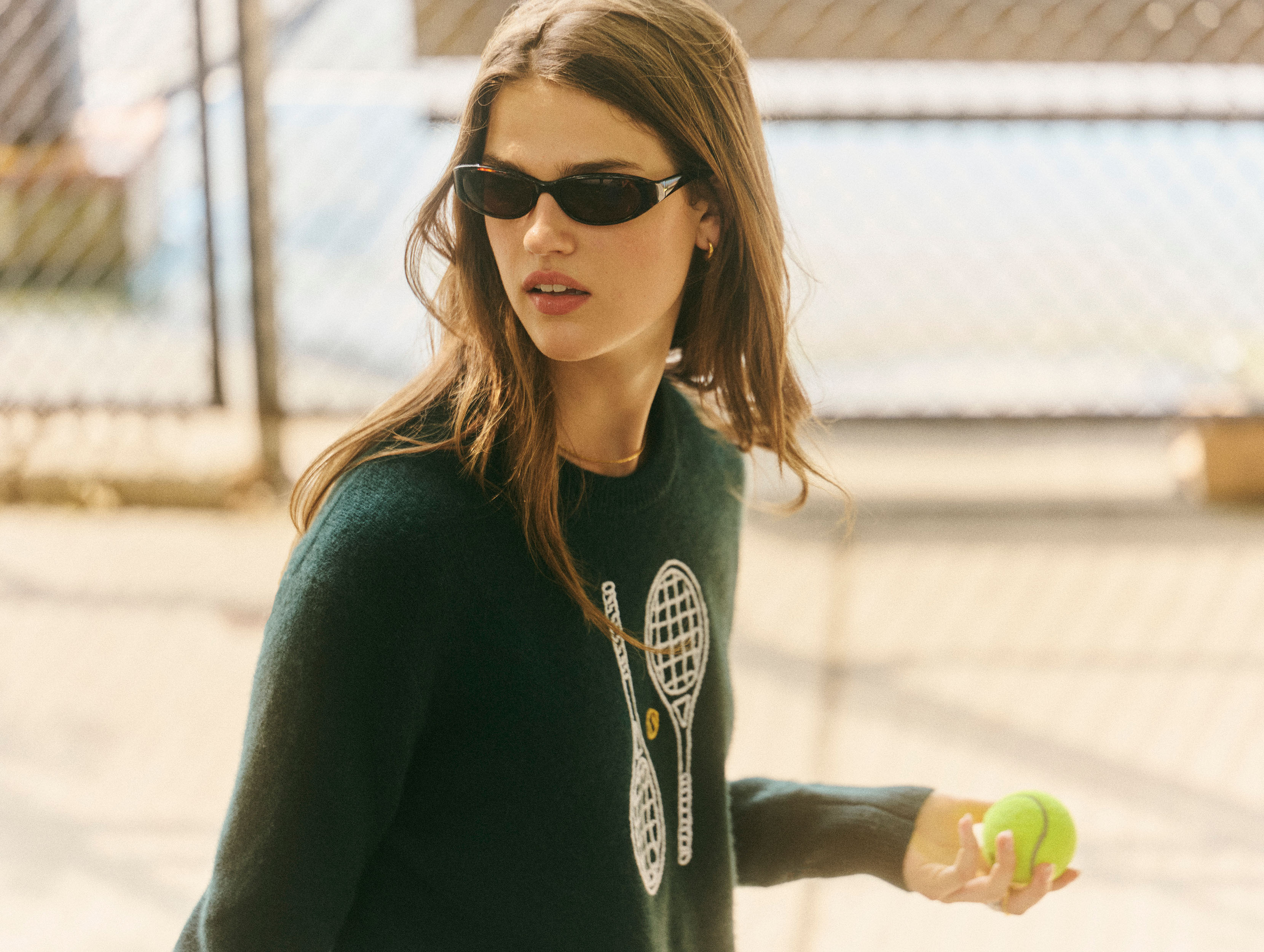 A woman wearing dark sunglasses and a dark green sweater with a tennis racket graphic, holding a tennis ball on an outdoor court.