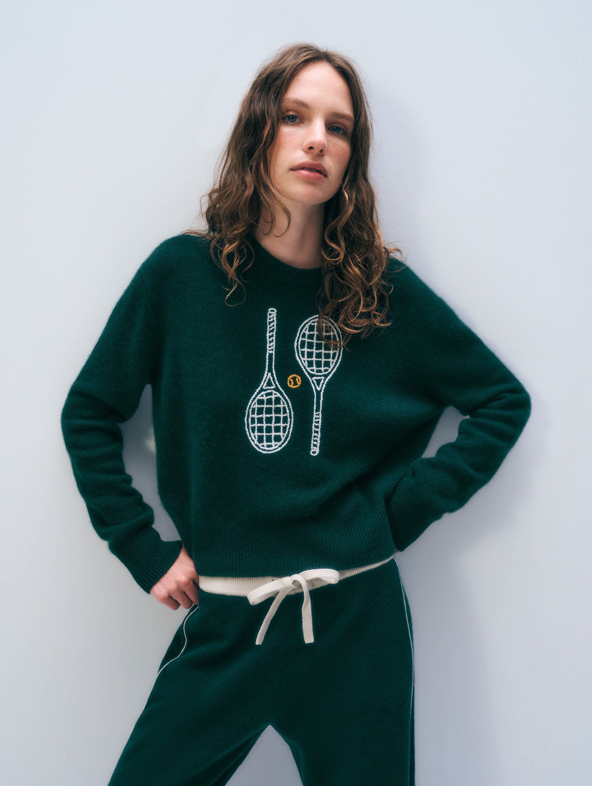 A woman wearing a dark green knit sweatshirt and matching sweatpants. The sweatshirt features a white graphic of two tennis rackets with a small yellow tennis ball motif in the center.