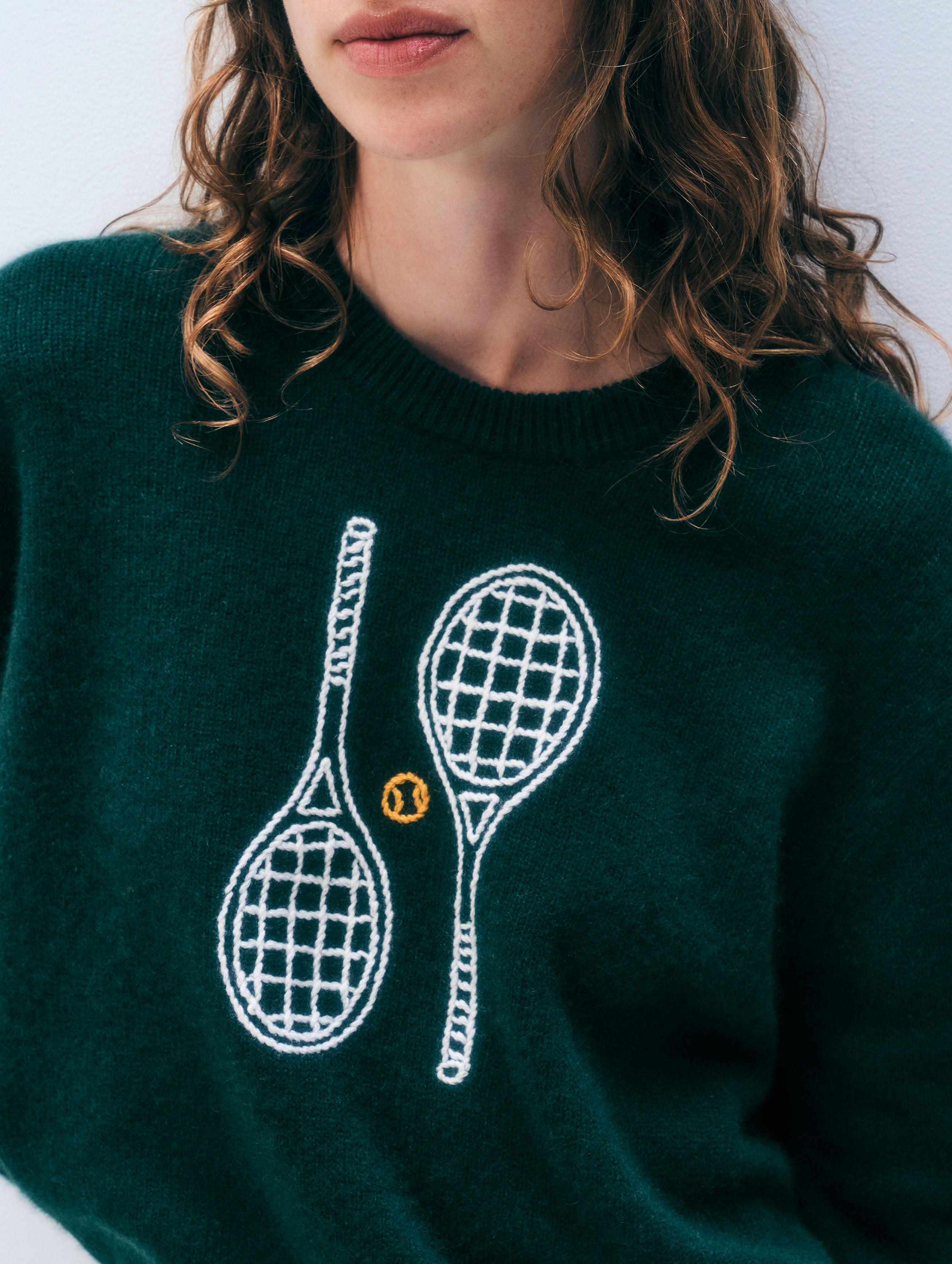 A close-up of a person wearing a dark green knit sweater with white embroidered tennis rackets and a small yellow tennis ball graphic on the front.