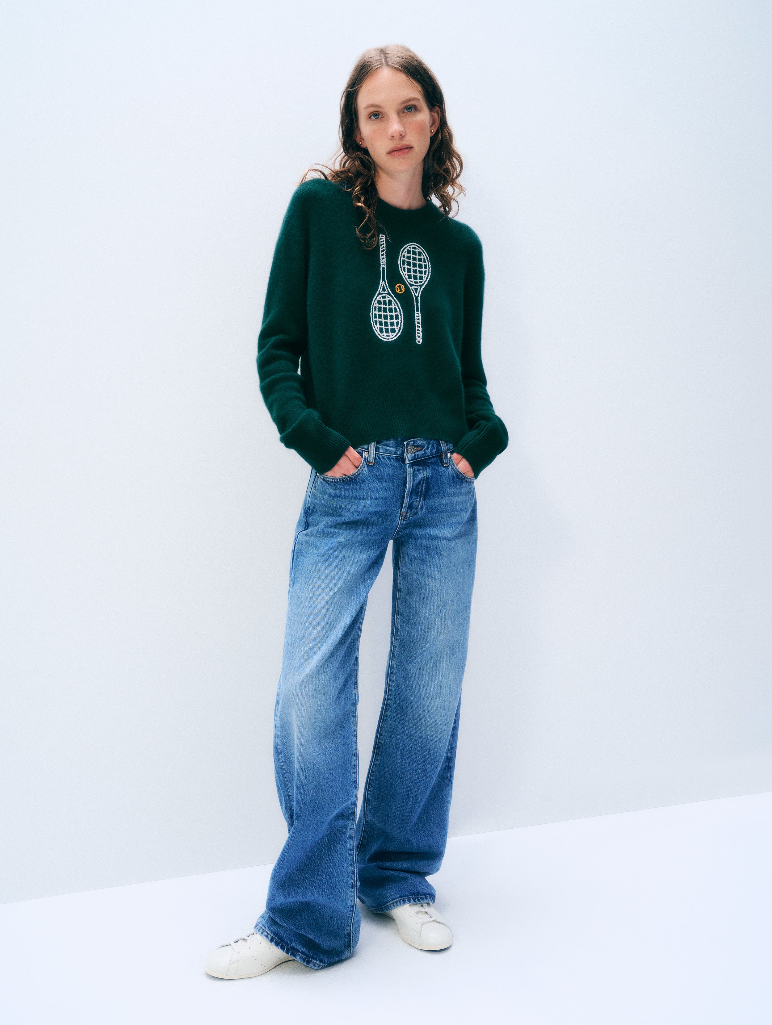 A woman stands against a plain light gray studio background wearing a dark green long-sleeve sweater with a tennis racket graphic on the front, blue wide-leg jeans, and white sneakers.