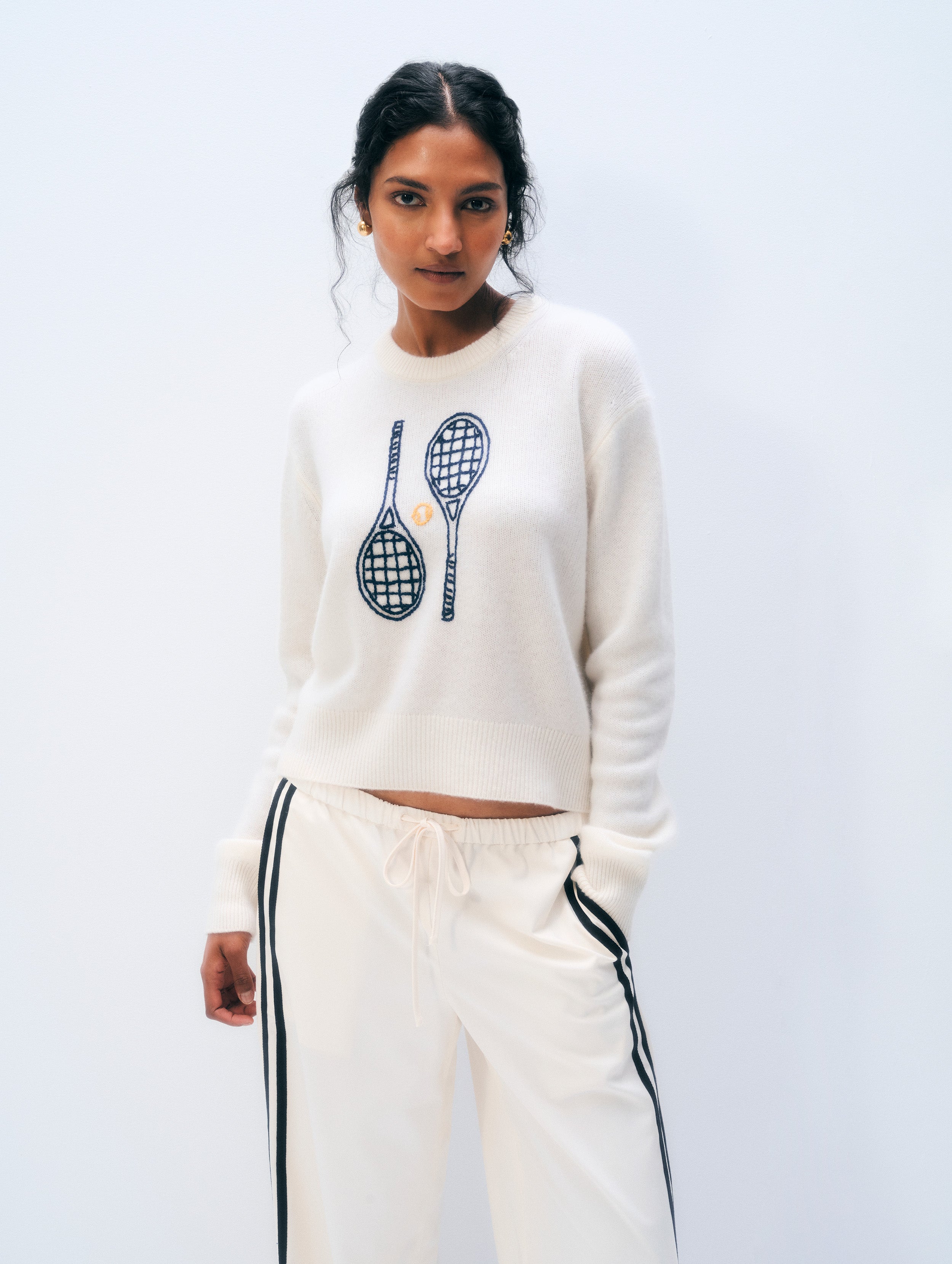A woman modeling a cream-colored cropped sweatshirt with a black tennis racket graphic on the chest, paired with matching cream track pants featuring black side stripes, against a plain light gray background.