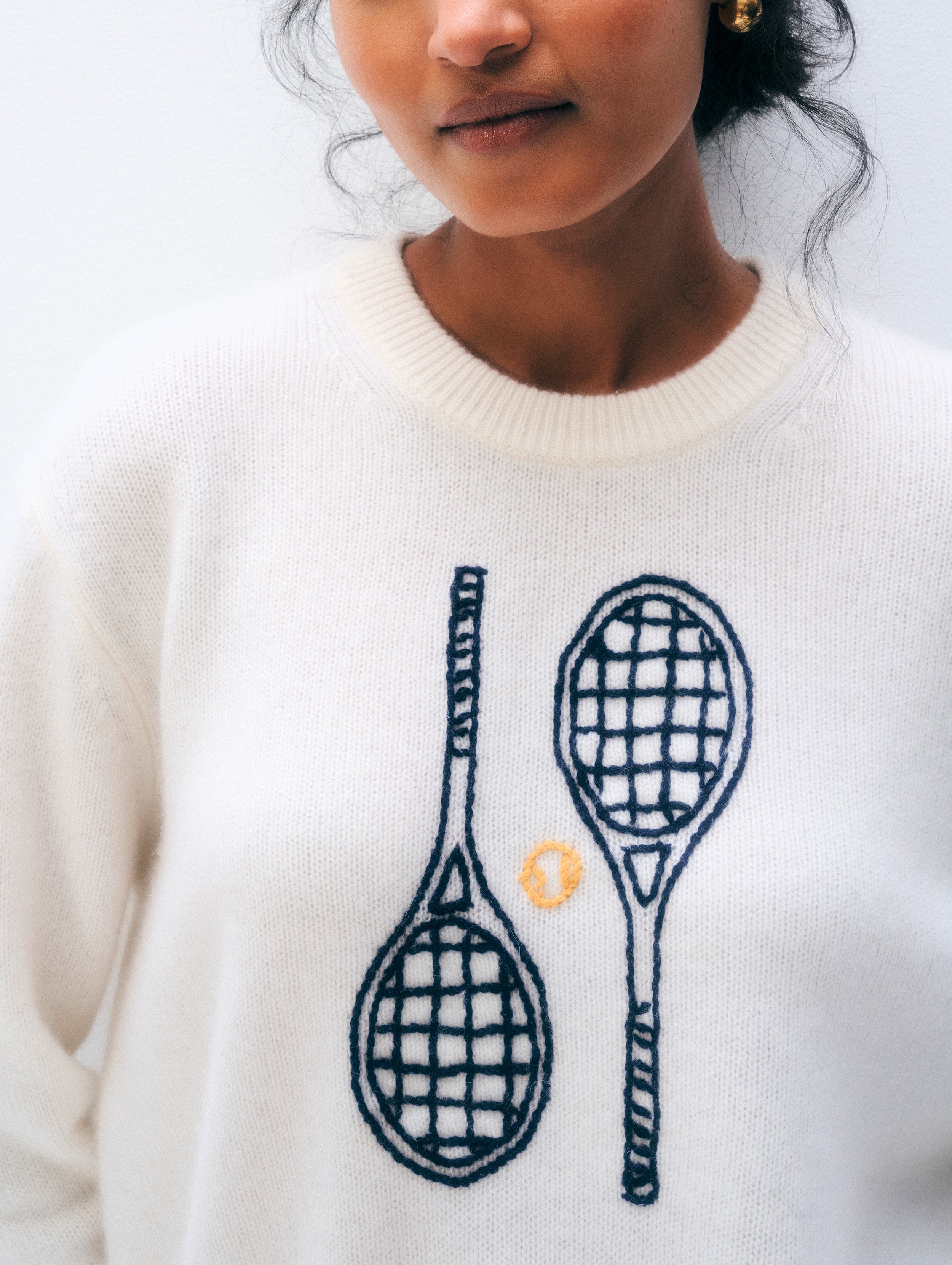 A close-up of a person wearing a cream-colored knit sweater with a graphic of two dark blue tennis rackets and a small yellow tennis ball on the front.