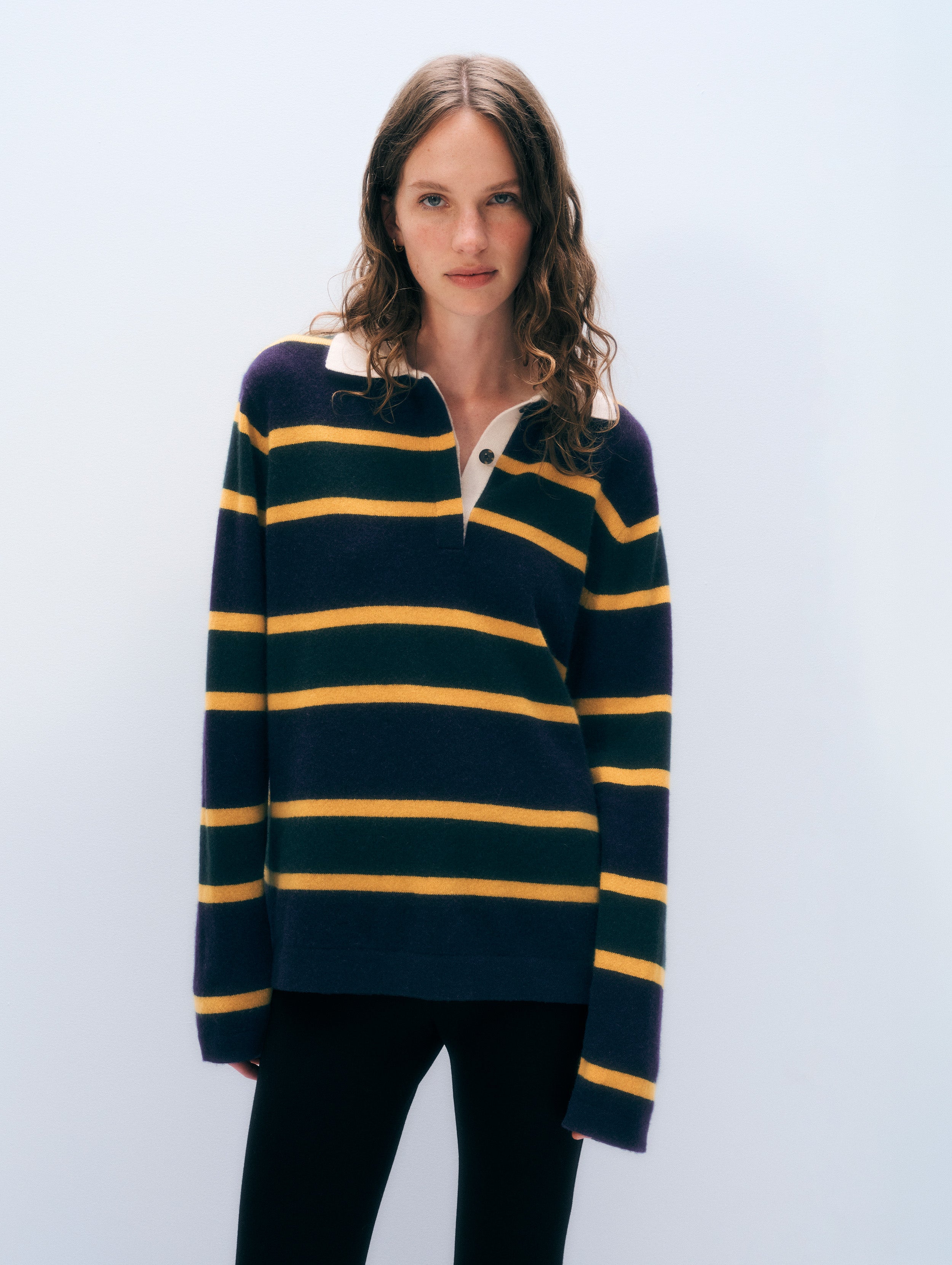 A woman modeling a navy blue and yellow striped rugby-style sweater with a white collar, paired with black pants, against a light gray studio background.