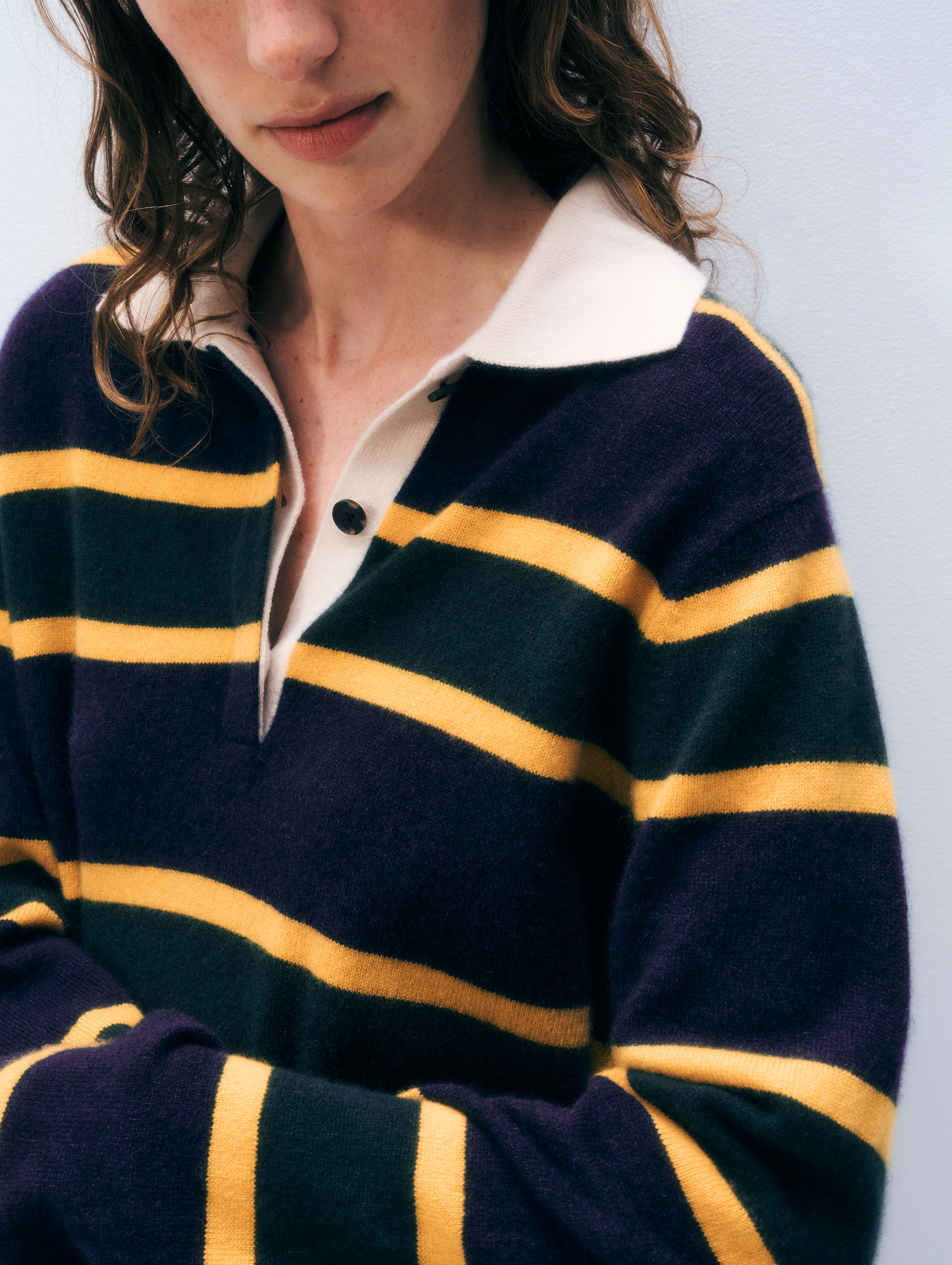 A close-up of a person wearing a knit rugby-style sweater with horizontal navy and mustard yellow stripes, a cream collar, and a partial button placket.