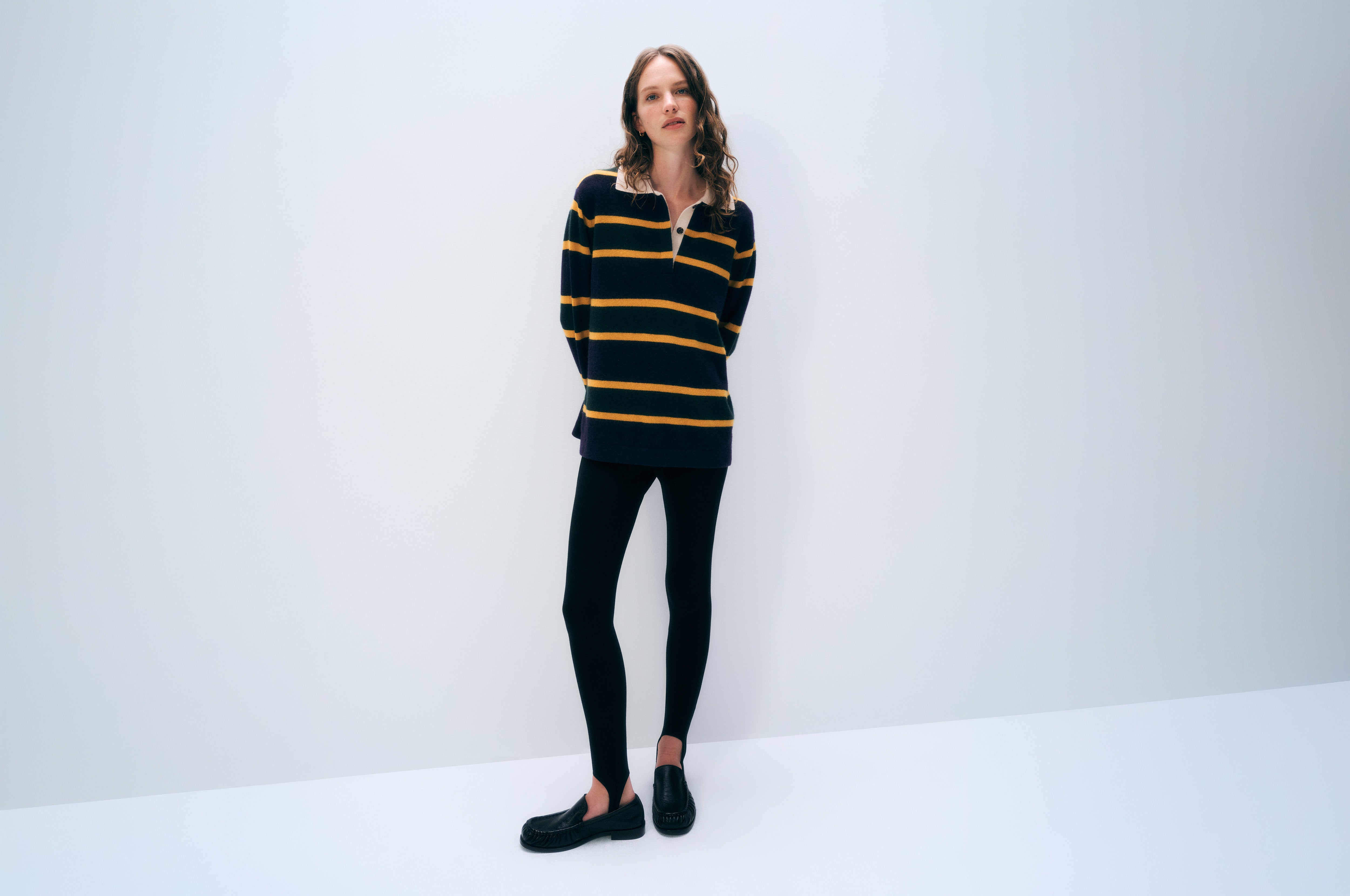 A woman stands against a plain white studio background wearing a black long-sleeve knit top with mustard yellow horizontal stripes, black leggings, and black slip-on shoes.