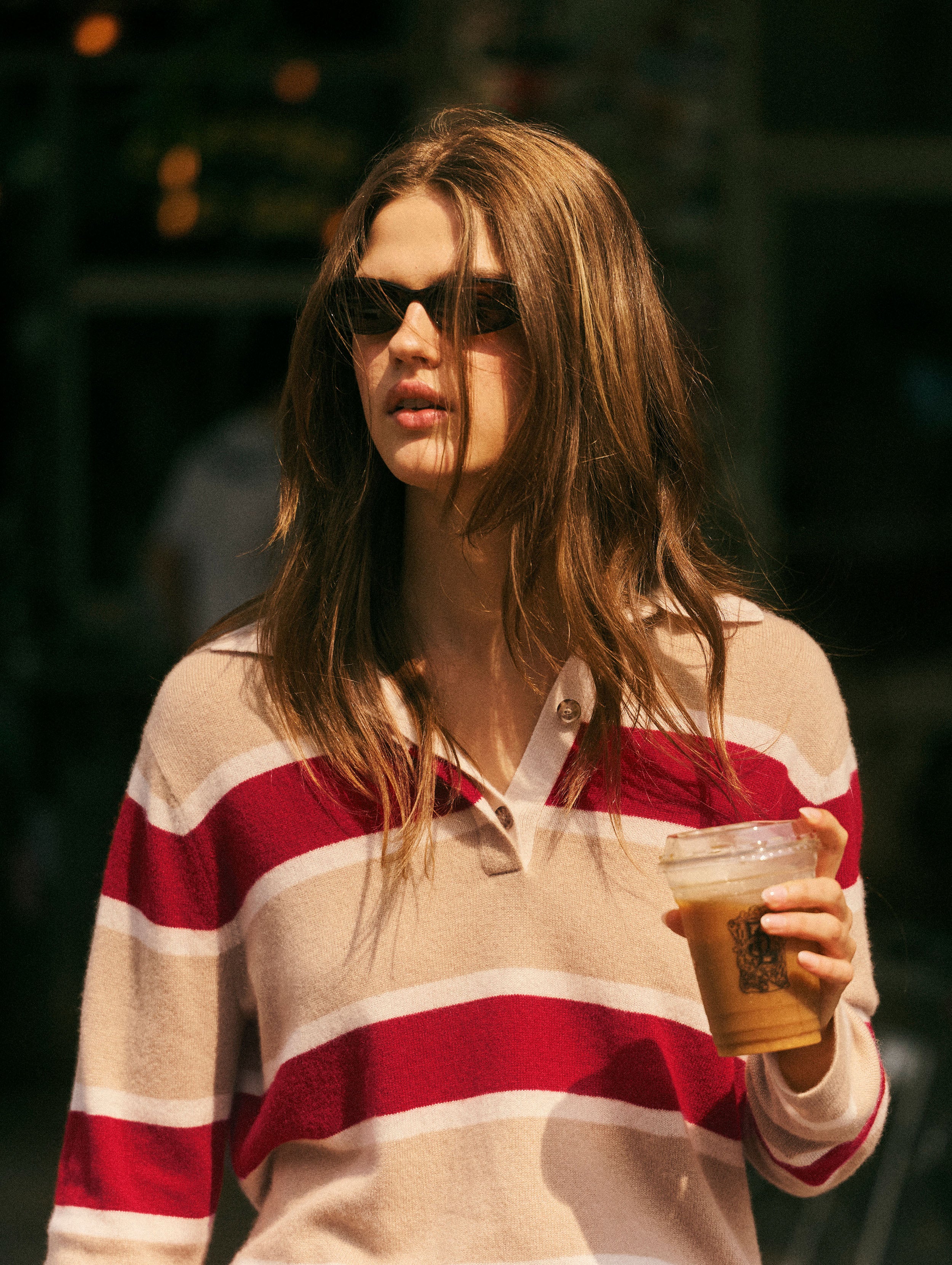 A woman wearing a beige and red striped knit polo sweater, dark sunglasses, and holding an iced coffee in a warm, outdoor setting.