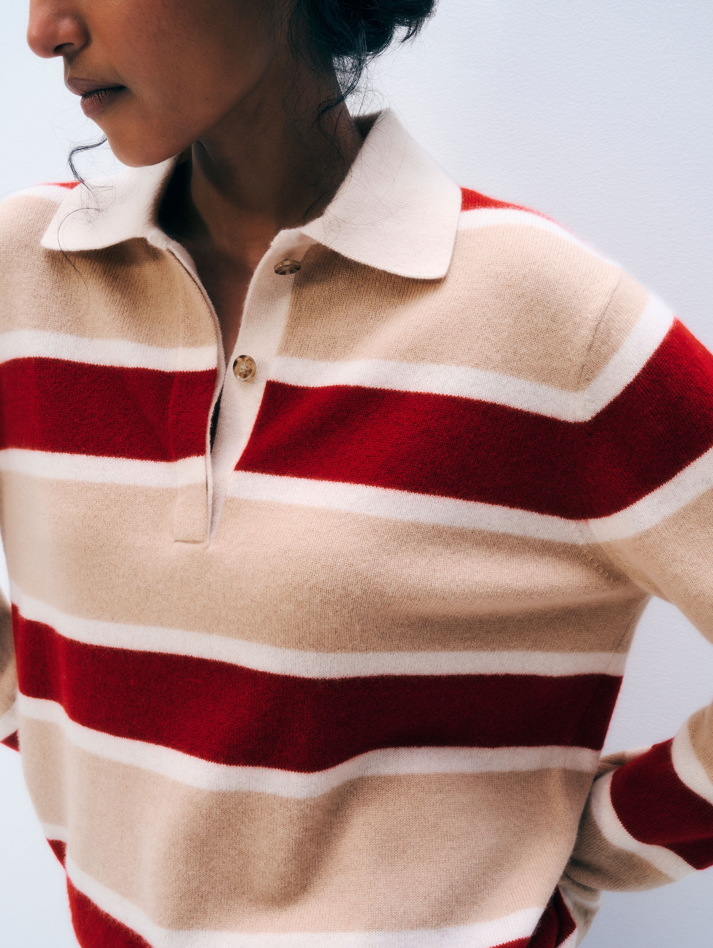 A close-up of a person wearing a knit polo sweater with horizontal stripes in beige, red, and white, featuring a white collar and a partial button placket.