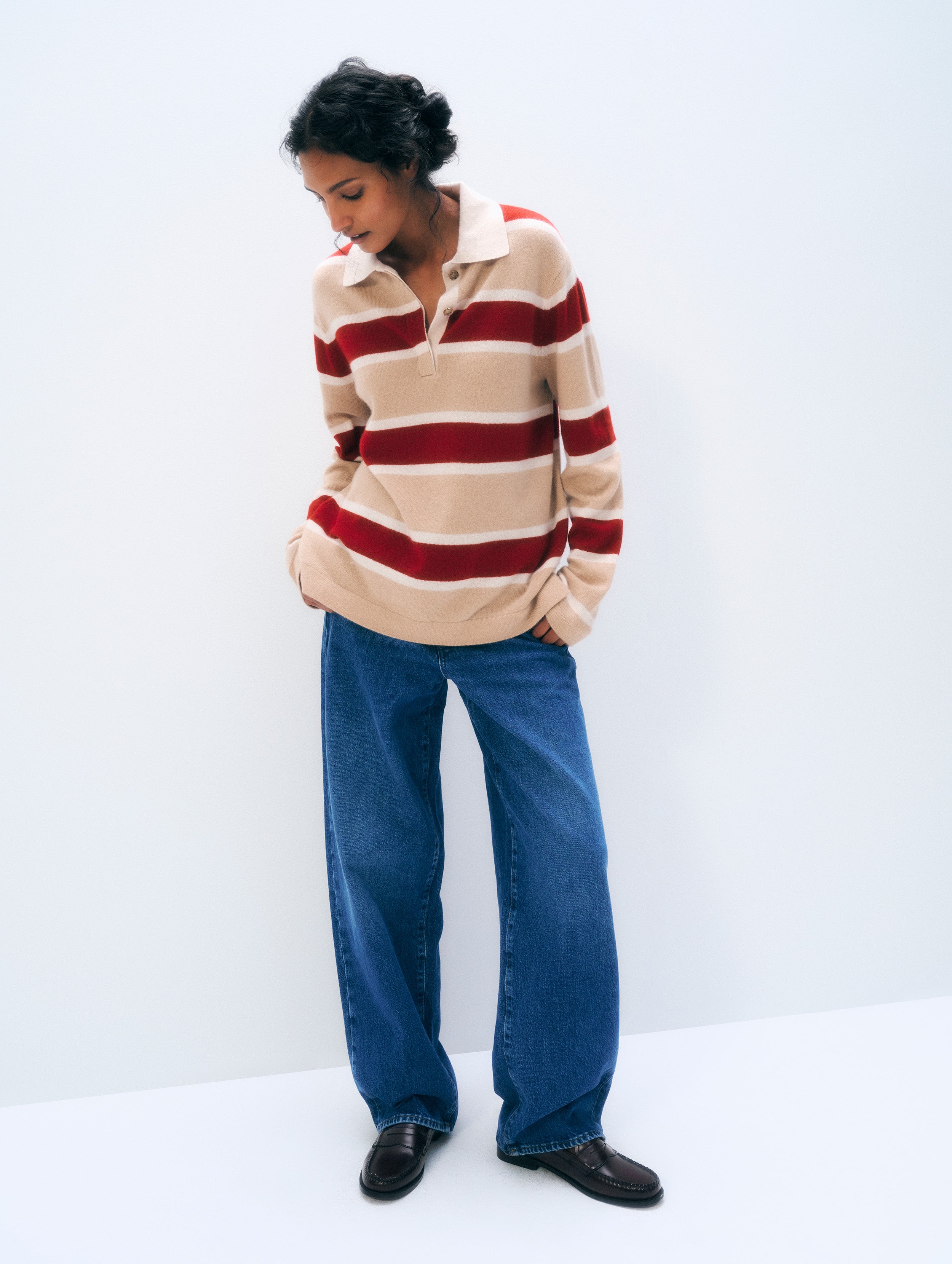 A woman wearing a beige and red striped long-sleeve polo sweater, wide-leg blue jeans, and black shoes, standing against a plain white studio background.
