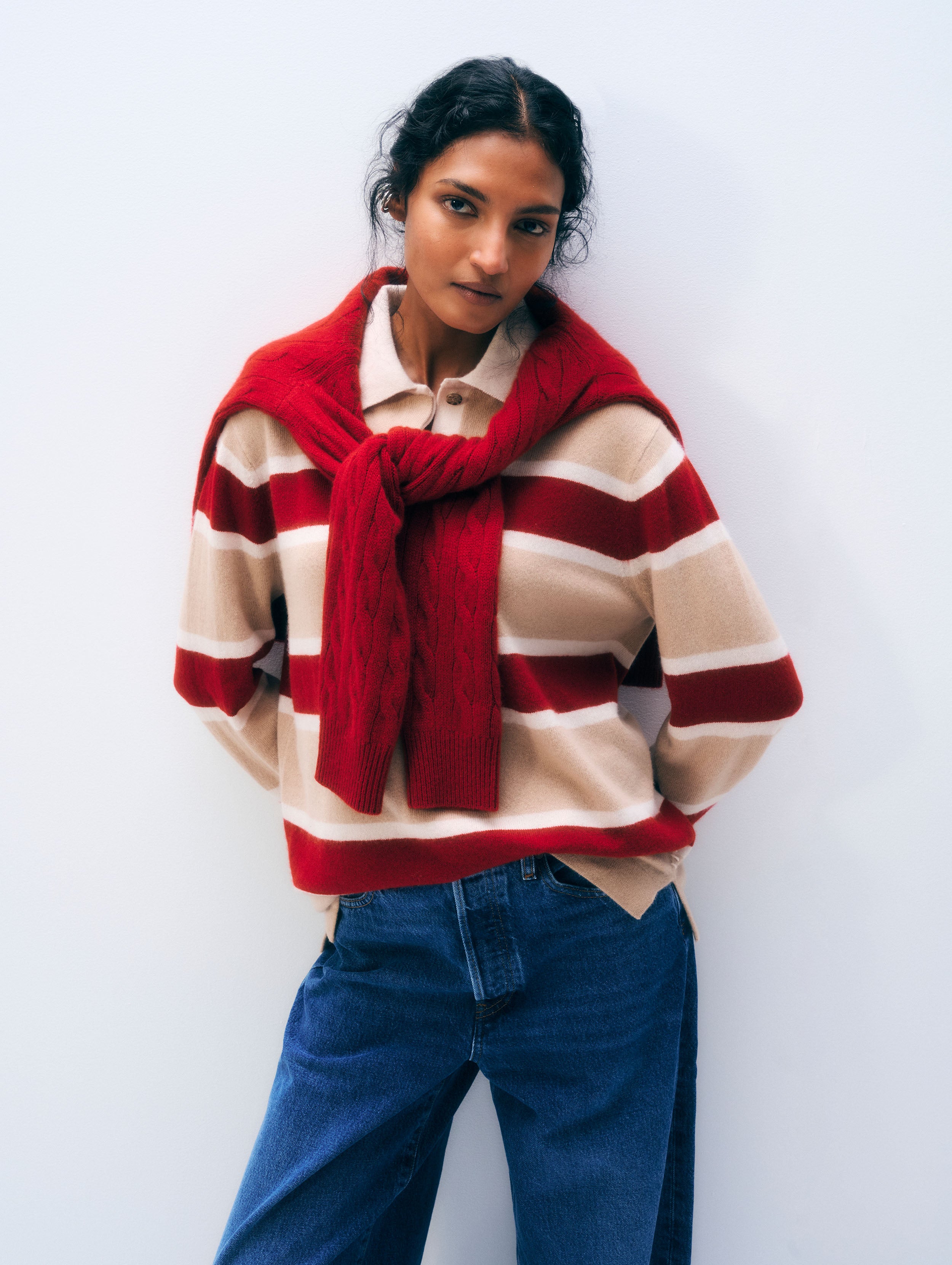 A woman wearing a cream and red striped knit sweater with a red knit scarf wrapped around her shoulders, paired with blue jeans, standing against a plain light gray wall.