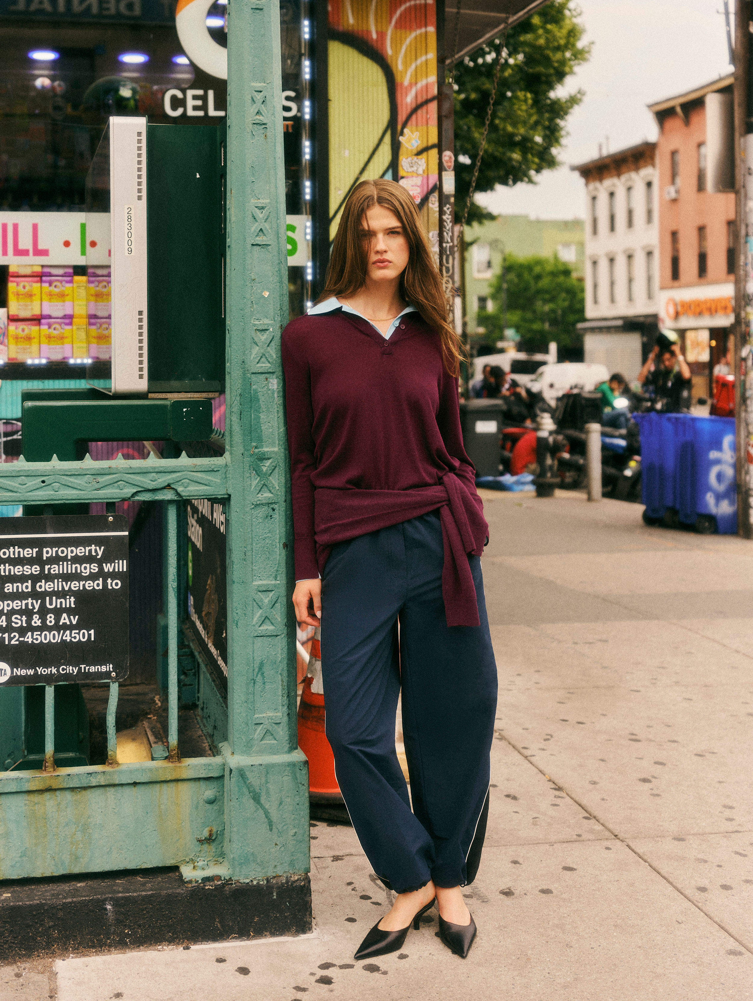 A woman stands on a city sidewalk wearing a burgundy knit sweater tied at the waist over a light blue collared shirt, navy pants, and black pointed flats. She is posed next to a green storefront column in an urban street setting.